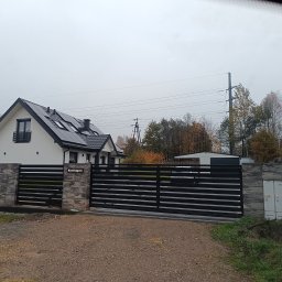 Bartłomiej Monica OGRO-TECH - Nowoczesna, przesuwna brama metalowa w kolorze czarnym, z poziomymi panelami, zamontowana przy posesji z białym domem i szarą kostką. Widok z zewnątrz.