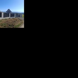 Bartłomiej Monica OGRO-TECH - Nowoczesne ogrodzenie panelowe z betonowymi słupkami na nierównym terenie. W tle dom i góry pod błękitnym niebem. Widok z boku, w słoneczny dzień.