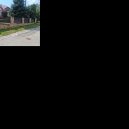 Bartłomiej Monica OGRO-TECH - Nowoczesne ogrodzenie z kamienną podmurówką i poziomymi, ciemnymi deskami, brama wjazdowa, dom w tle, trawnik i chodnik. Ujęcie z poziomu ulicy.