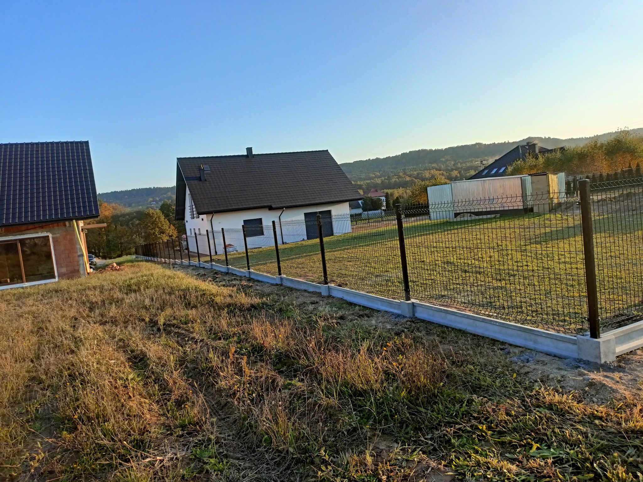 Nowoczesne ogrodzenie panelowe z podmurówką betonową na nierównym terenie, w tle domy jednorodzinne i zalesione wzgórza w słoneczny dzień. Widok z dołu.