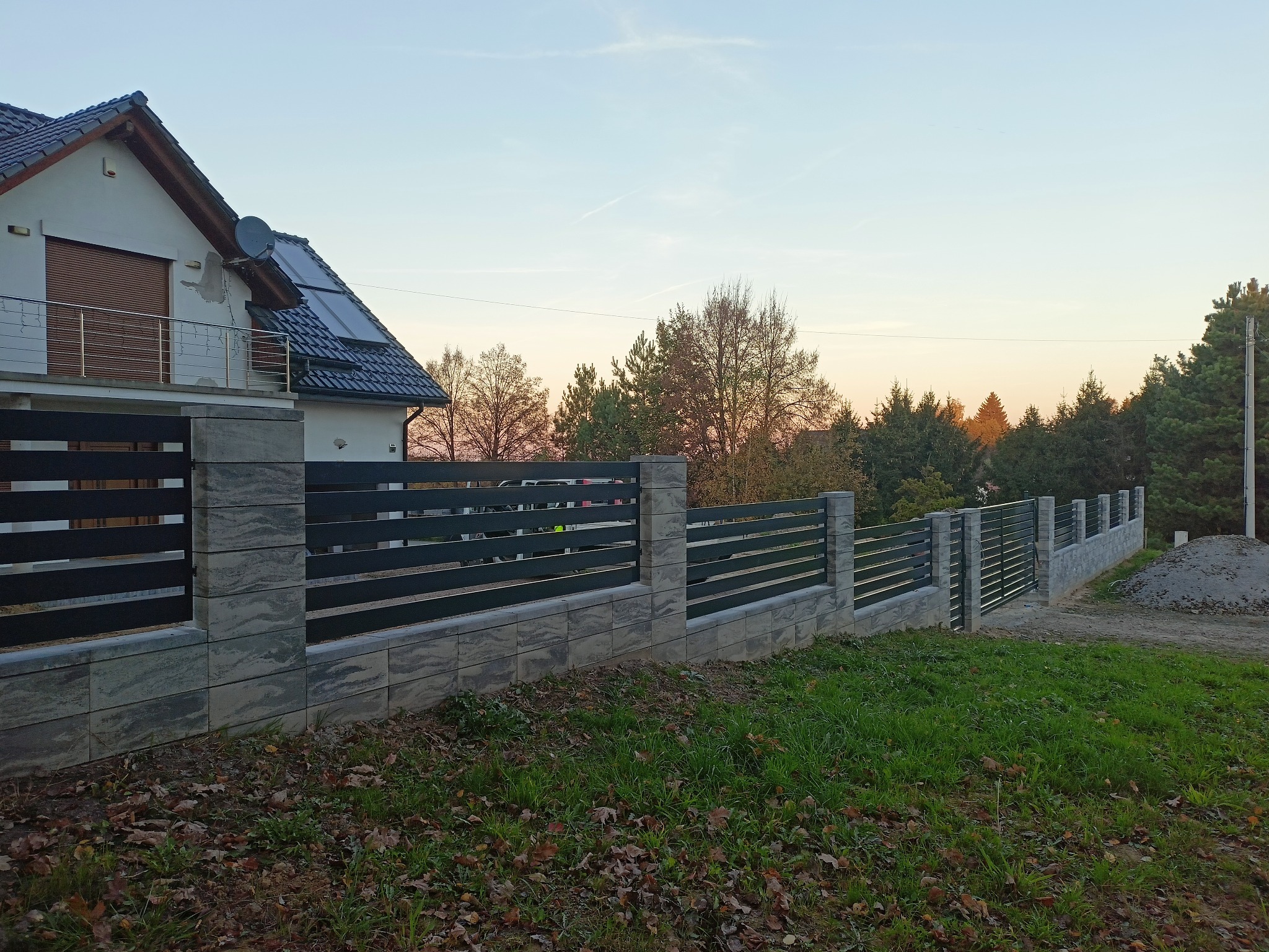 Nowoczesne ogrodzenie panelowe z elementami betonowymi, widoczne na tle domu jednorodzinnego o zmierzchu. Charakterystyczne poziome panele i solidne słupki.