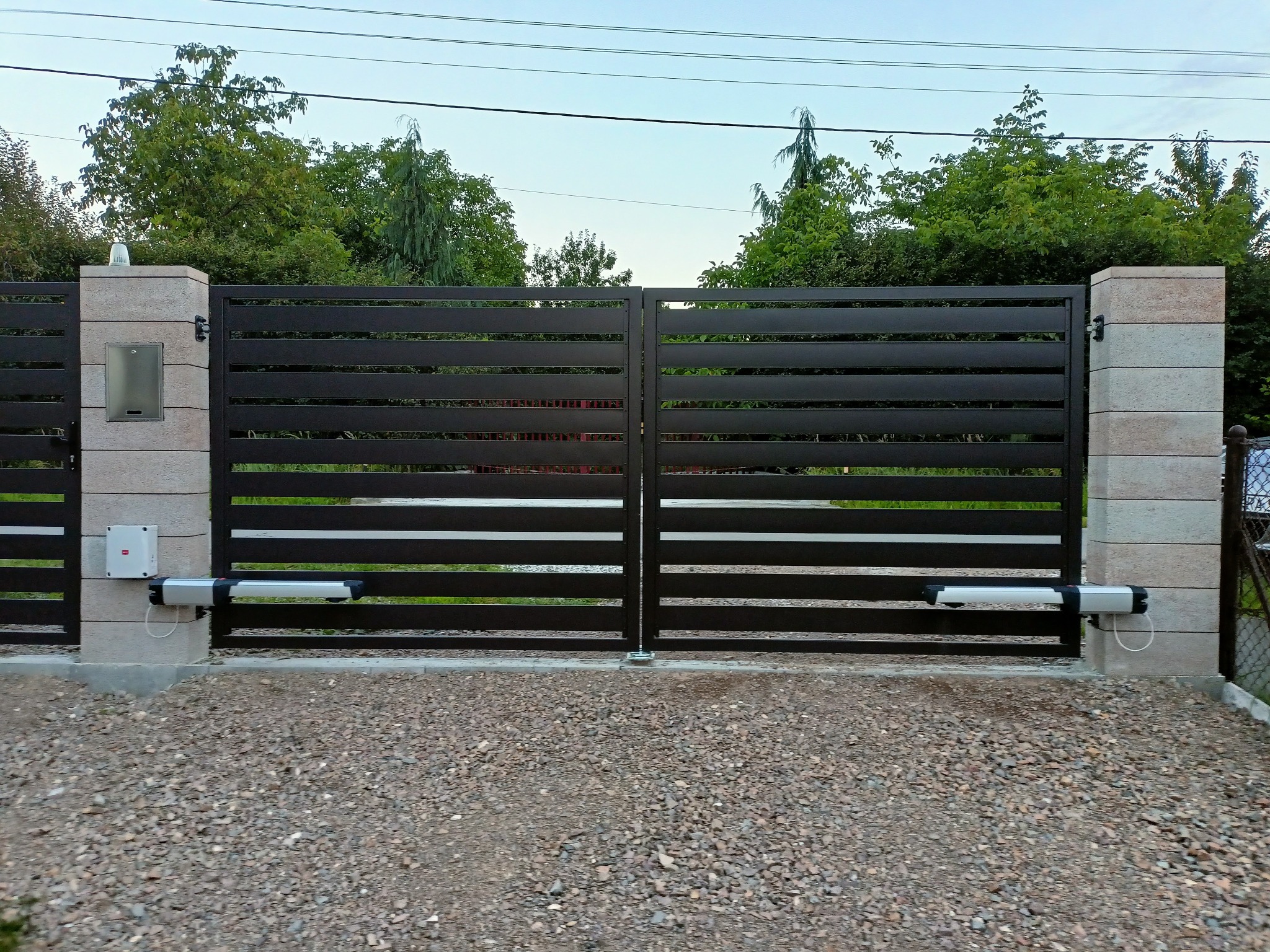 Nowoczesna brama przesuwna z automatyką, poziome lamele w kolorze grafitowym, osadzona na słupkach z imitacji kamienia. Widoczne siłowniki i skrzynka na listy.
