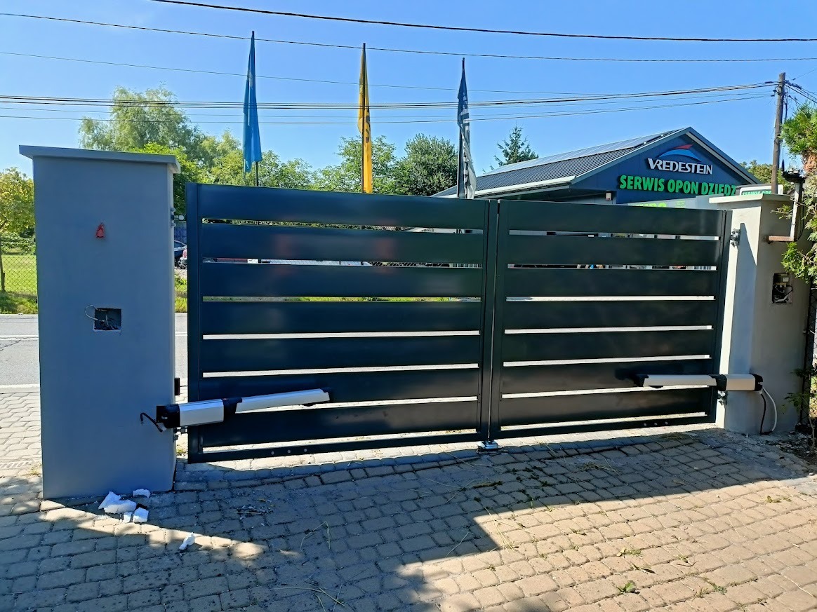 Nowoczesna, pozioma brama metalowa w kolorze grafitowym z automatyką, zamontowana na betonowych słupkach. Widoczne okablowanie i fragmenty styropianu po montażu.