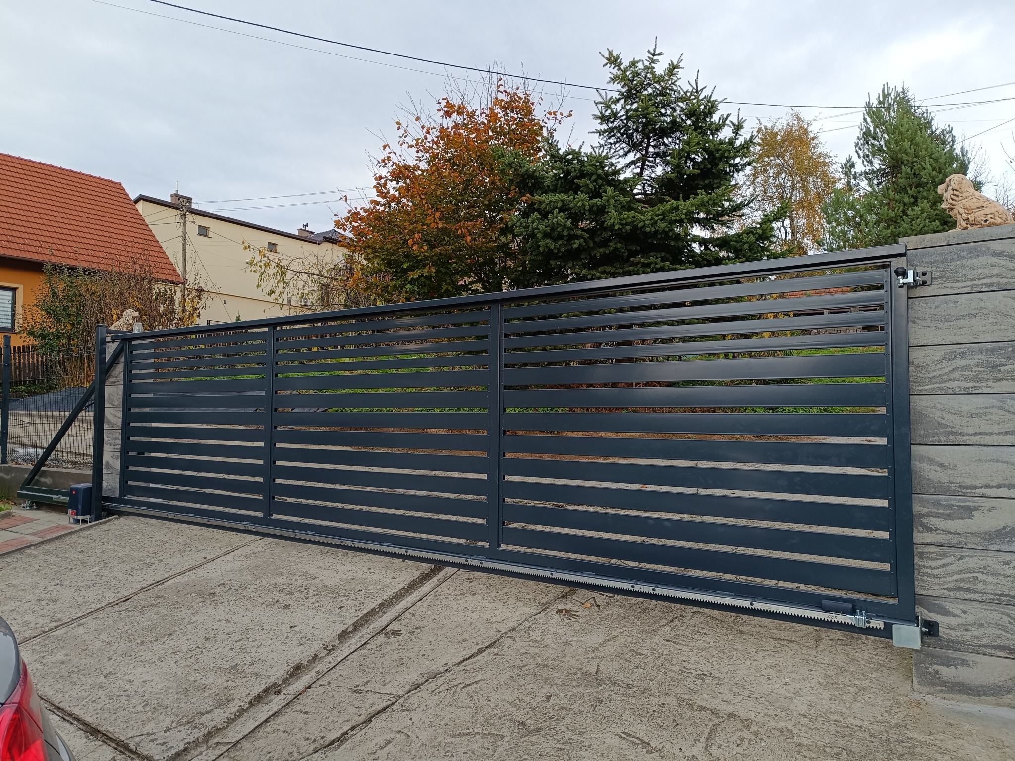 Nowoczesna, przesuwna brama w kolorze antracytu z automatyką, zamontowana na posesji z betonowym ogrodzeniem i rzeźbą lwa. Widok z boku, jesienne drzewa w tle.