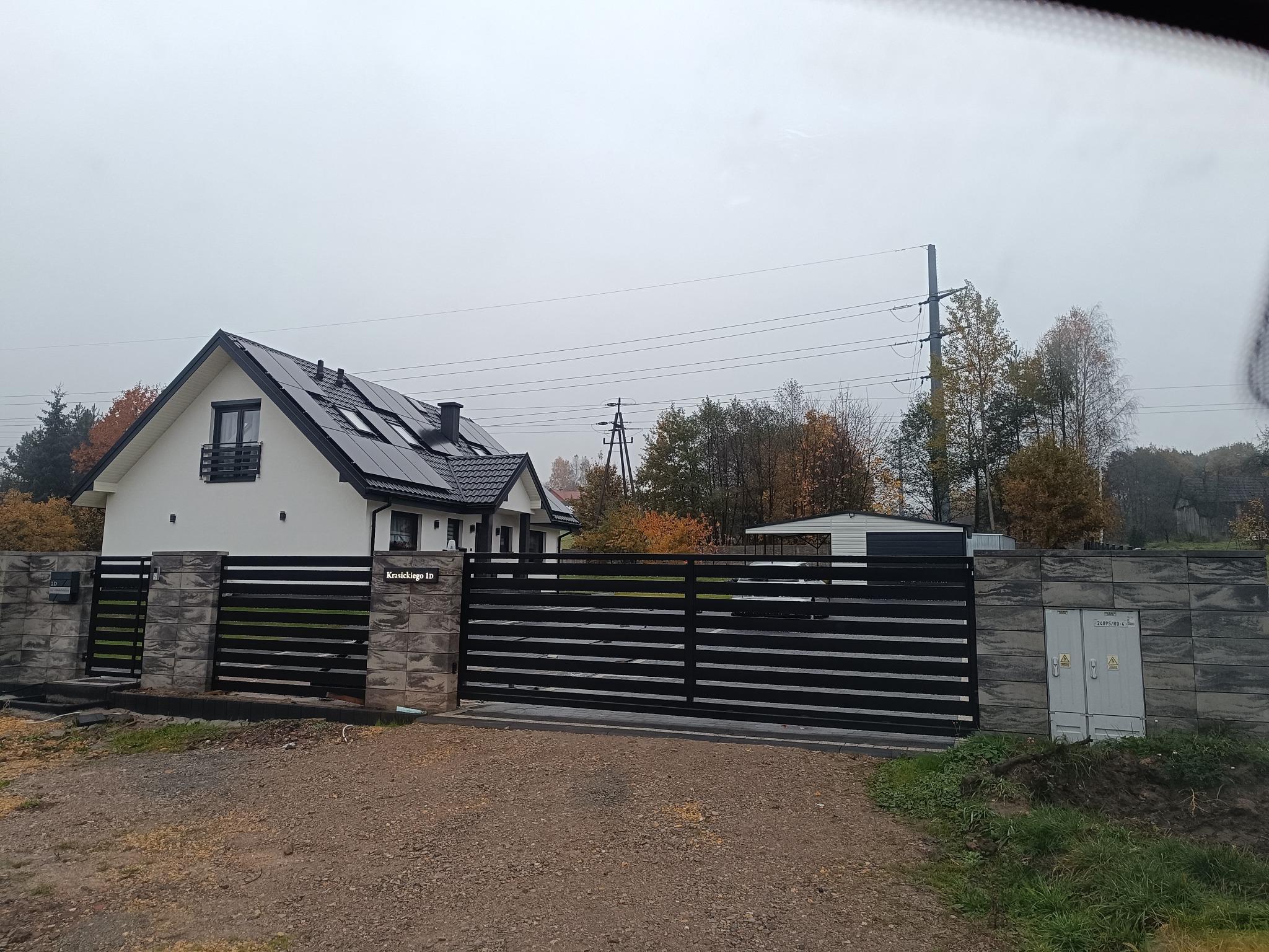 Nowoczesna, przesuwna brama metalowa w kolorze czarnym, z poziomymi panelami, zamontowana przy posesji z białym domem i szarą kostką. Widok z zewnątrz.