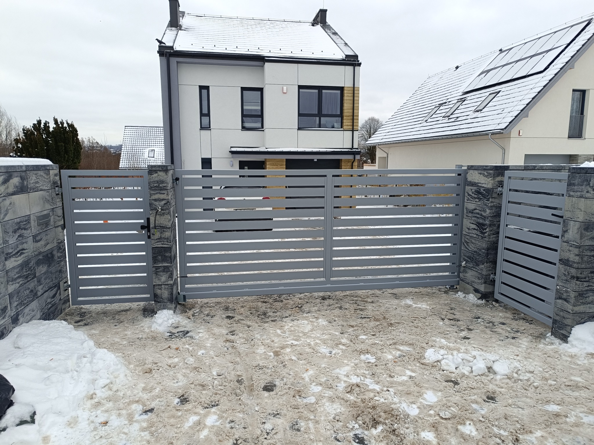 Nowoczesna, pozioma brama wjazdowa z szarego metalu, osadzona na kamiennych słupkach. W tle domy z dachami pokrytymi śniegiem. Zimowy krajobraz.