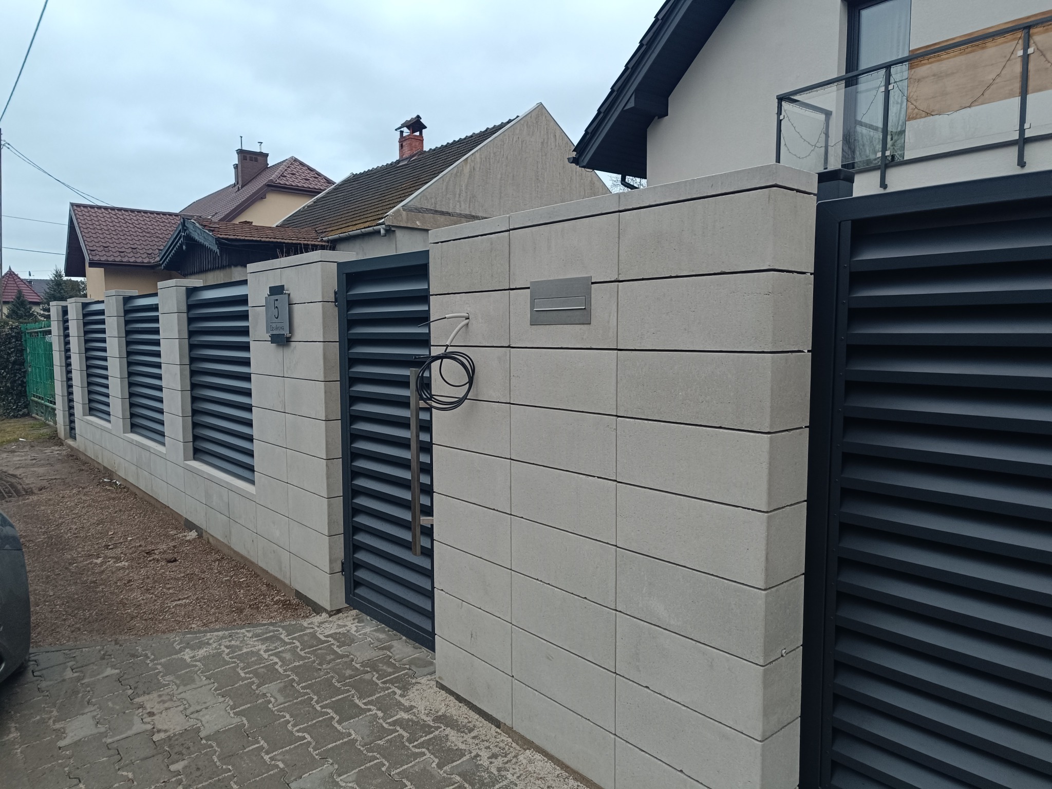 Nowoczesne ogrodzenie z szarego betonu i grafitowych paneli poziomych, z numerem domu i skrzynką pocztową. Widok z zewnątrz na posesję.