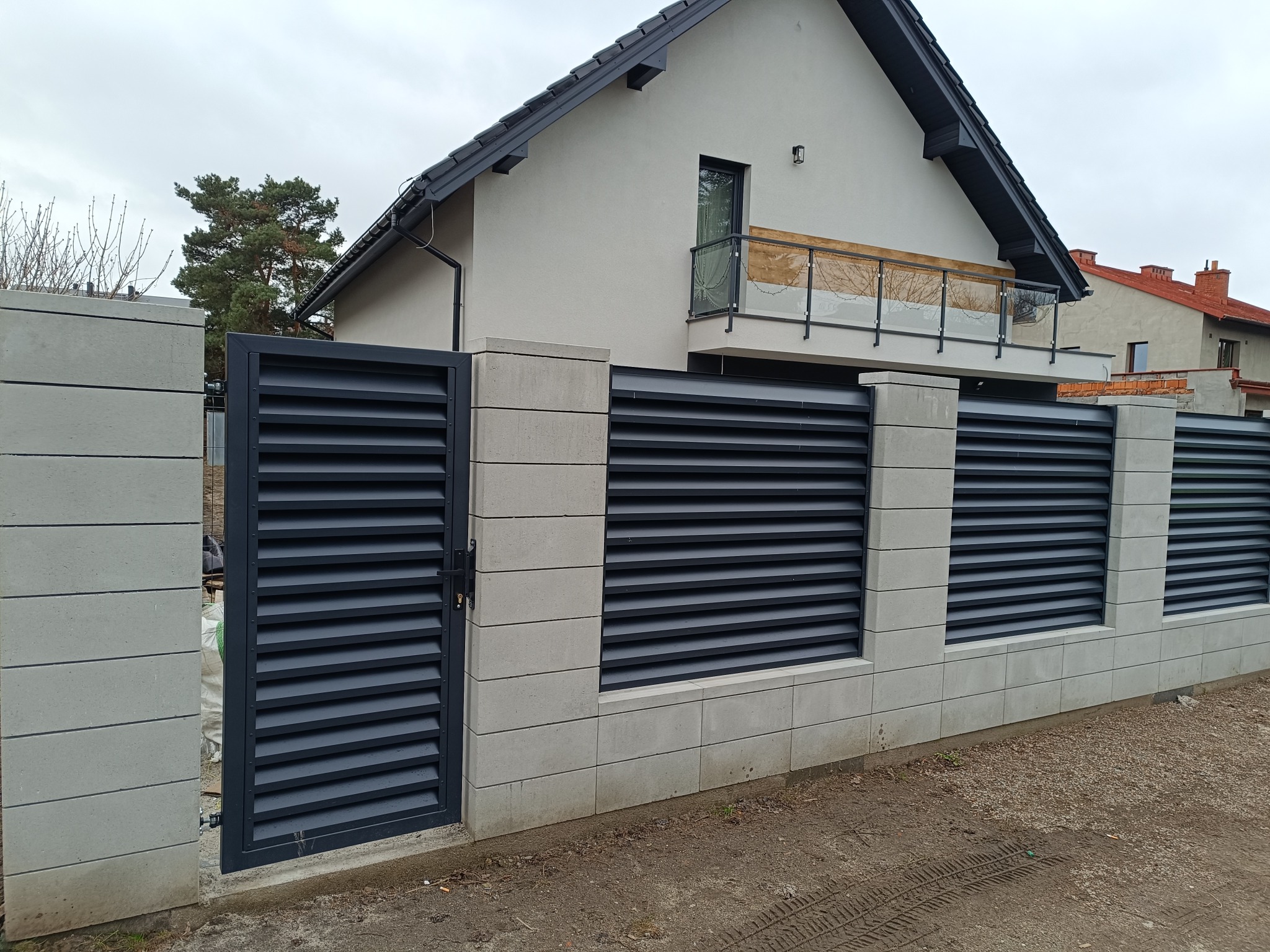 Nowoczesne ogrodzenie z szarych, betonowych bloczków i poziomych, grafitowych paneli. Brama lamelowa zamyka posesję z domem z balkonem w tle.