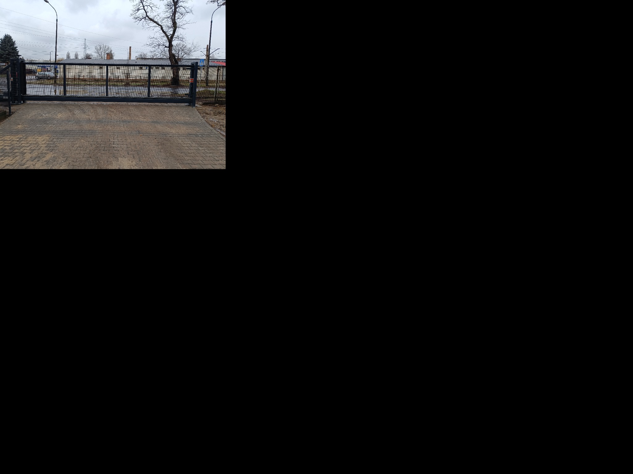 Metalowa brama przesuwna w kolorze grafitowym, z widoczną kostką brukową na podjeździe. W tle budynki przemysłowe i pochmurne niebo. Widok z zewnątrz posesji.