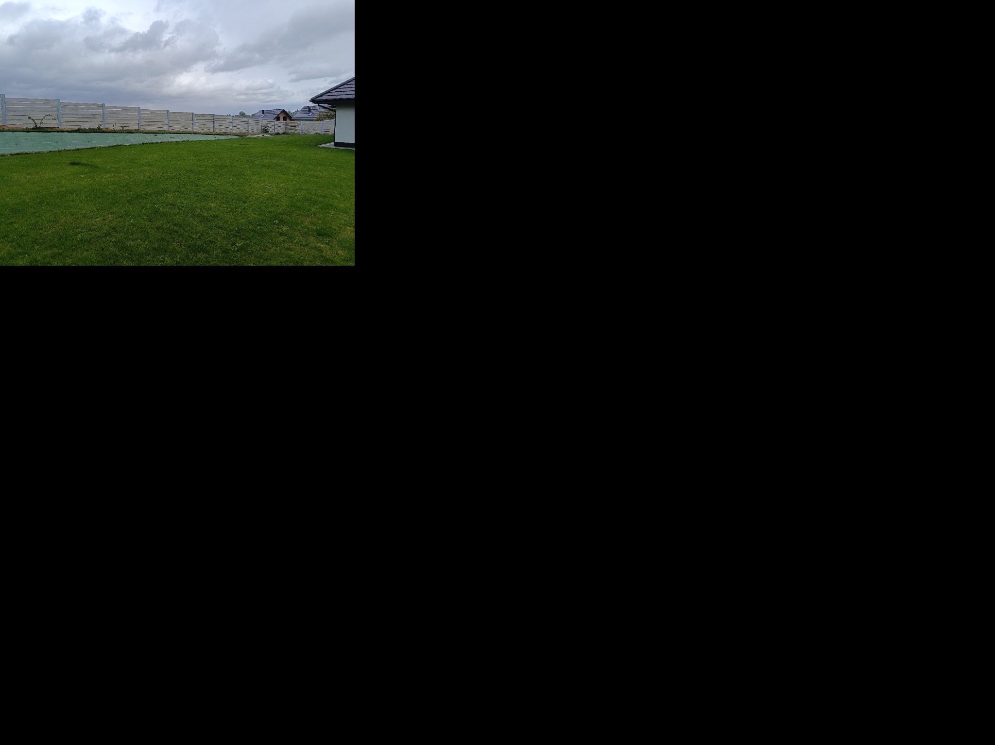 Długi, jasny płot betonowy, z widocznymi słupkami, otacza zielony trawnik przed domem. Pochmurne niebo w tle. Ujęcie z niskiej perspektywy.