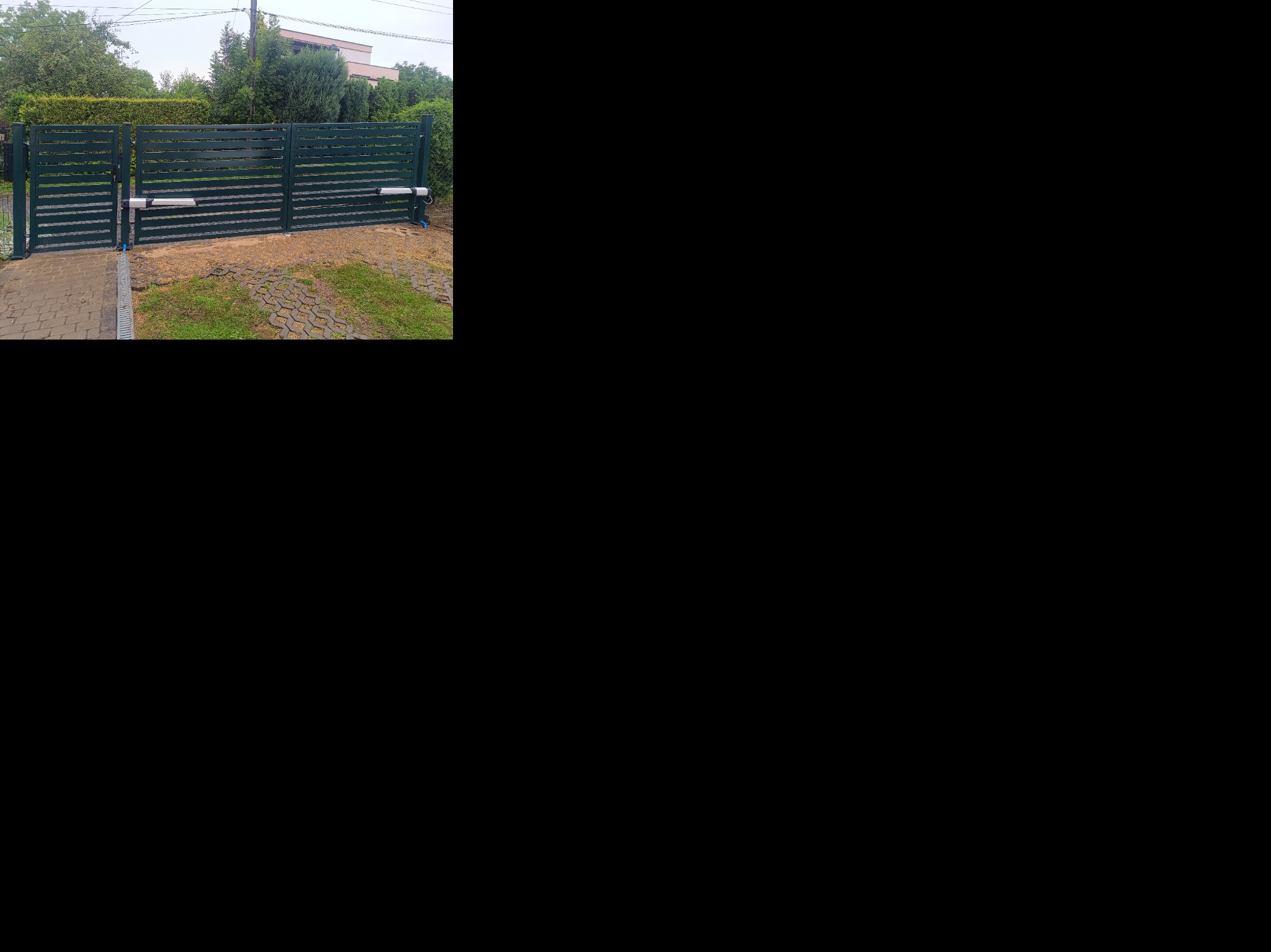 Nowoczesna, pozioma brama metalowa w kolorze ciemnej zieleni z automatyką, zamontowana na posesji z kostką brukową i zielenią w tle. Widoczne siłowniki bramy.