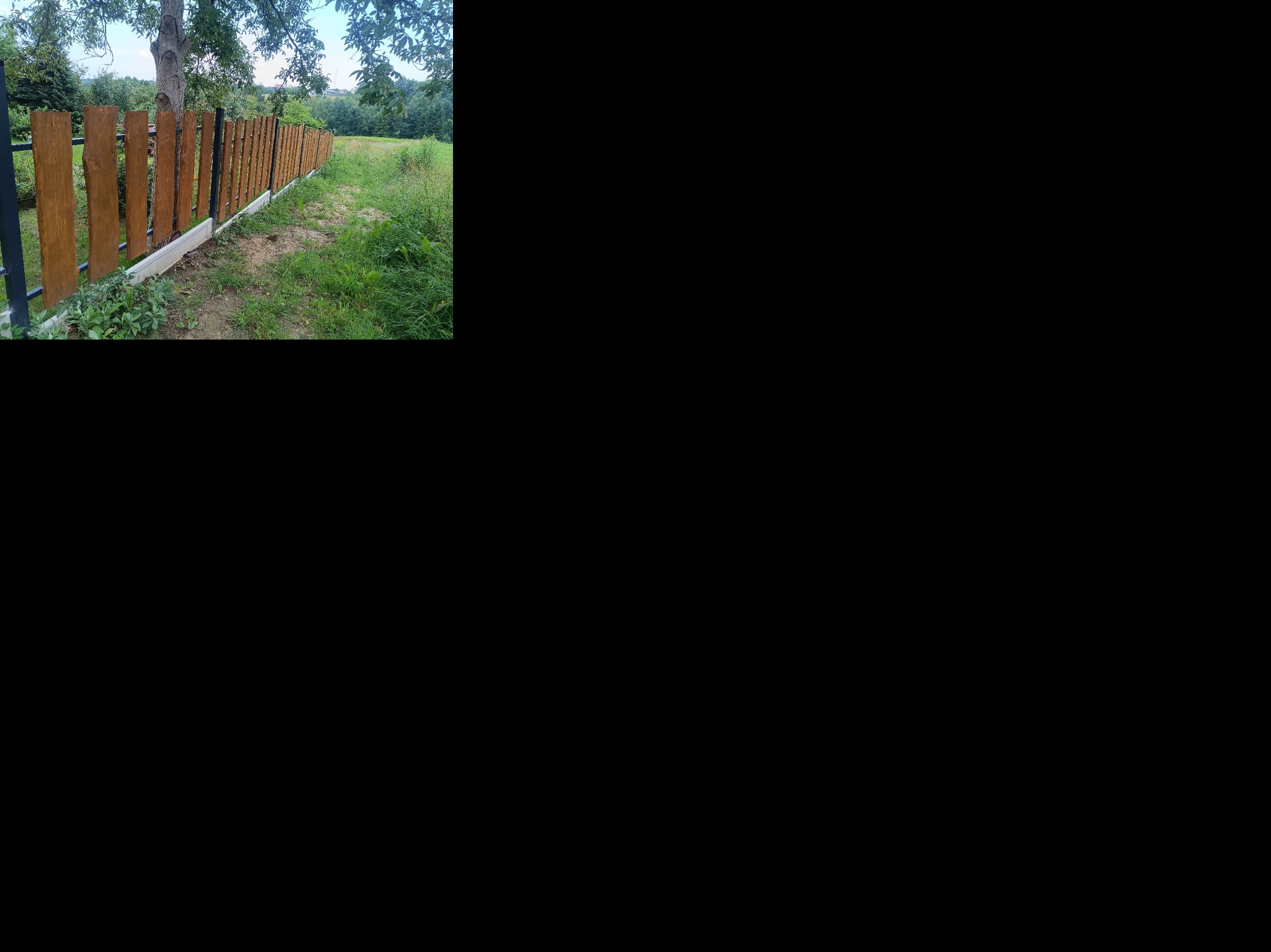 Drewniane ogrodzenie z metalowymi słupkami, osadzone na betonowej podmurówce, otoczone zielenią. Widok z perspektywy bocznej, w tle drzewa i trawa.