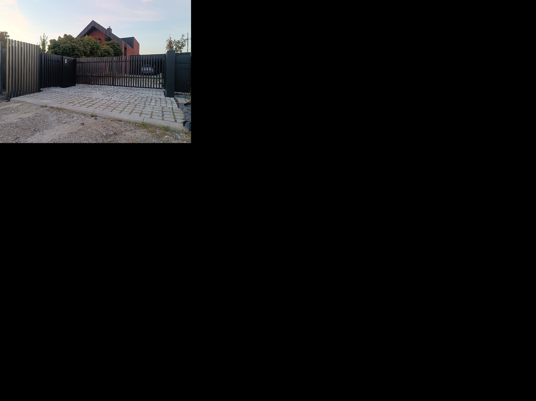 Nowoczesne, czarne ogrodzenie panelowe z bramą wjazdową na posesję z kostką. Widoczny fragment domu z czerwonej cegły w tle.