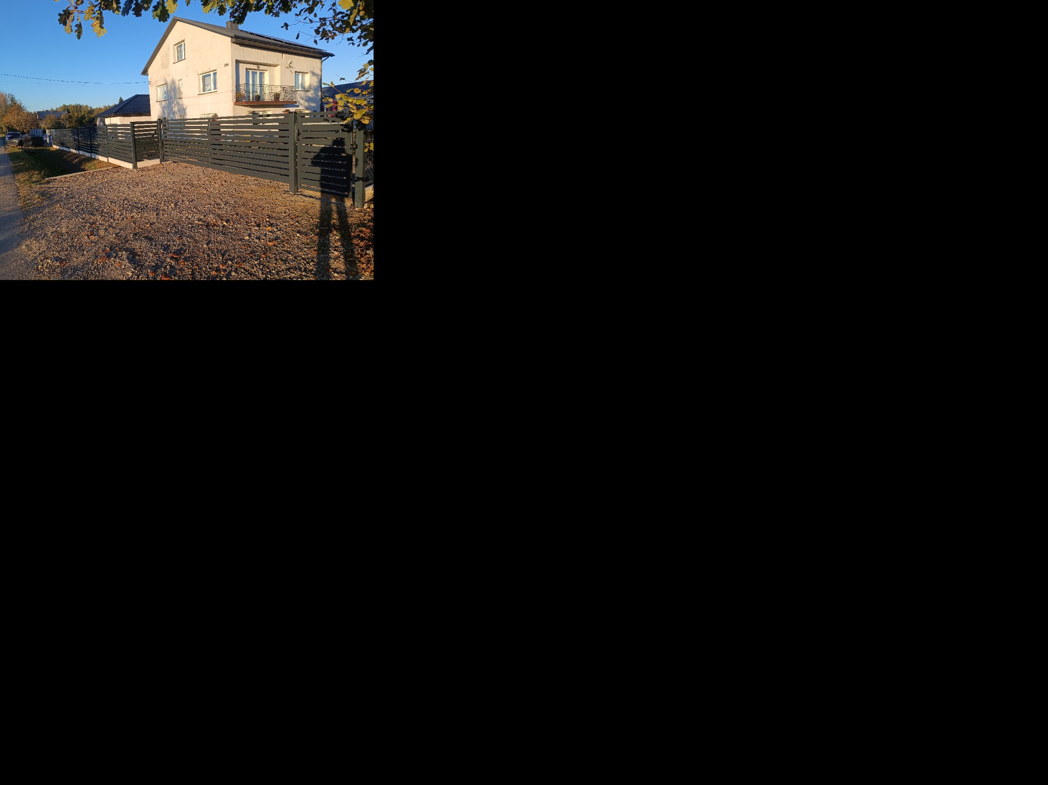 Nowoczesne, poziome ogrodzenie metalowe w kolorze grafitowym, z bramą wjazdową, na tle domu z balkonem i żwirowego podjazdu w słoneczny dzień.