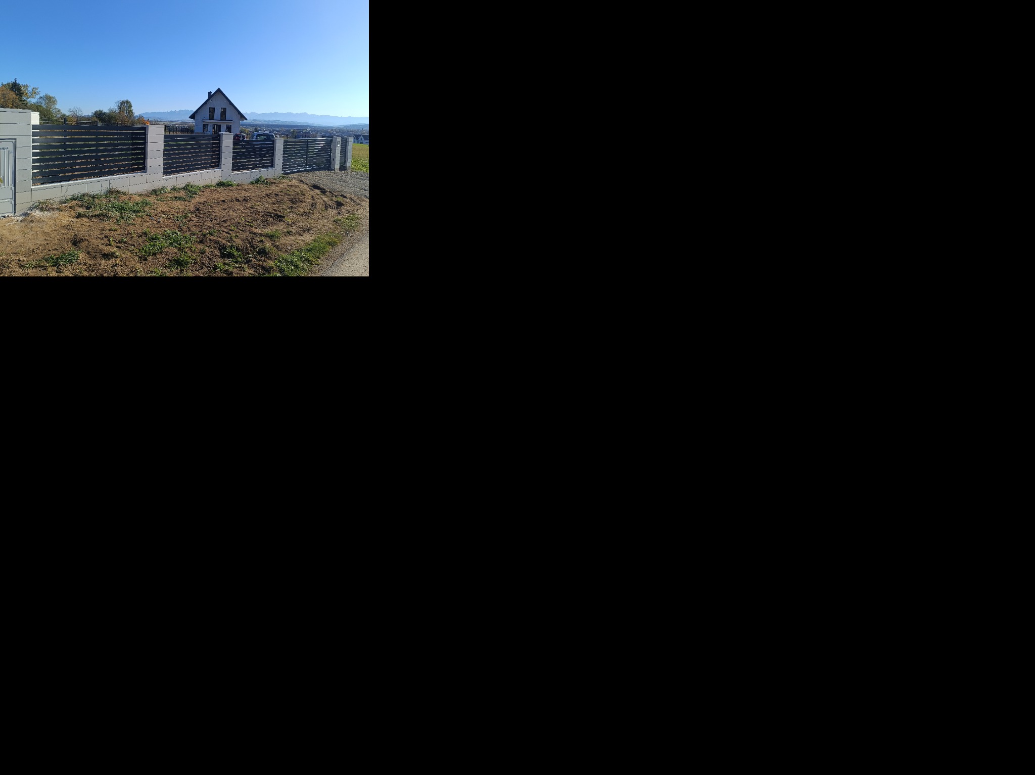 Nowoczesne ogrodzenie panelowe z betonowymi słupkami na nierównym terenie. W tle dom i góry pod błękitnym niebem. Widok z boku, w słoneczny dzień.