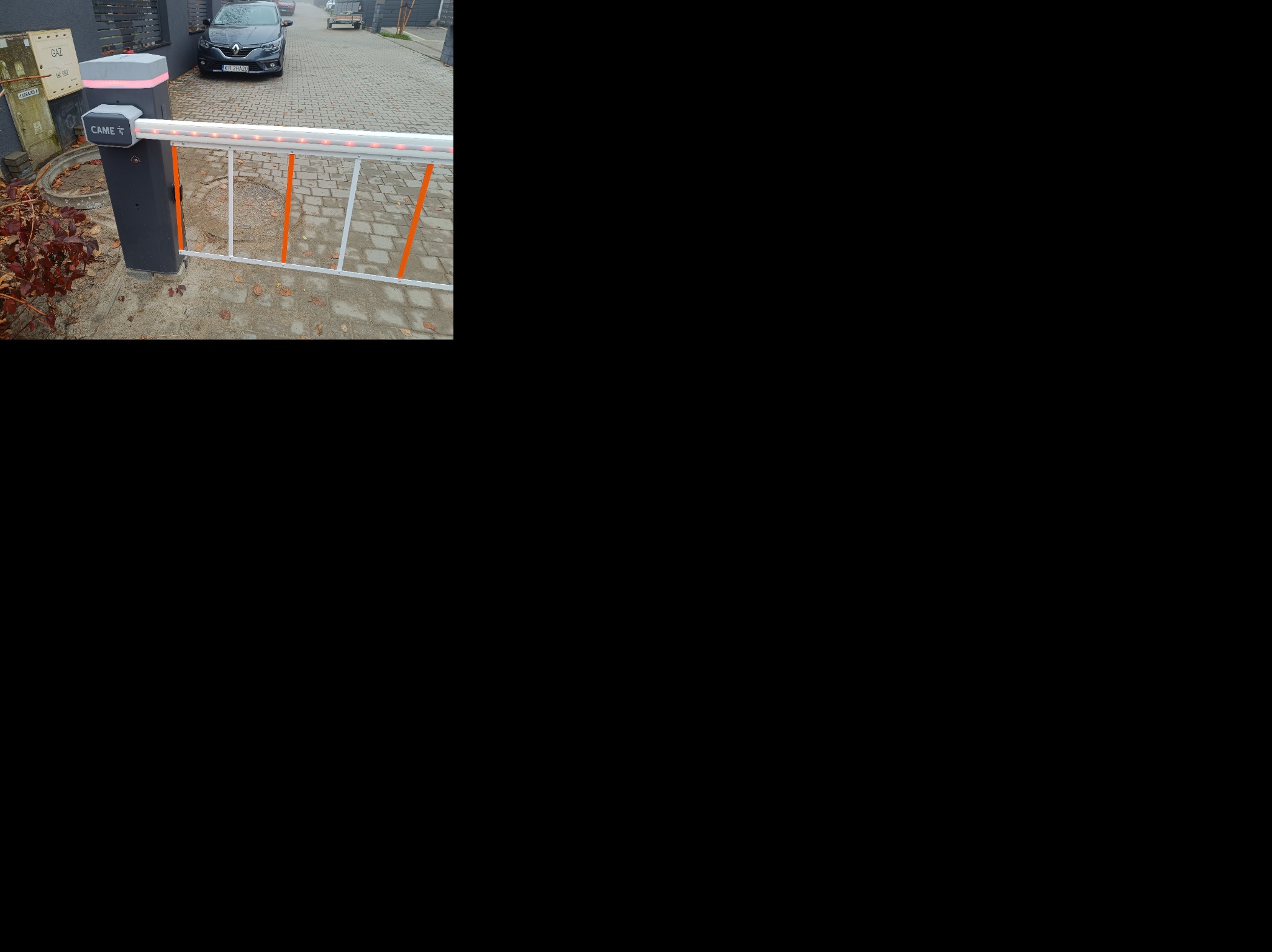 Szary szlaban automatyczny CAME z pomarańczowymi akcentami, zamontowany przy posesji z kostką brukową i zaparkowanym autem. Widoczny element skrzynki gazowej.