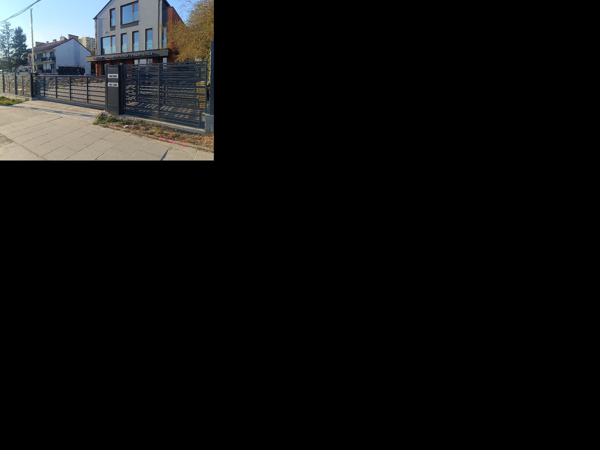 Nowoczesne, poziome ogrodzenie metalowe w kolorze grafitowym, z przesuwną bramą i furtką, przed budynkiem z szarą elewacją. Widok z chodnika.