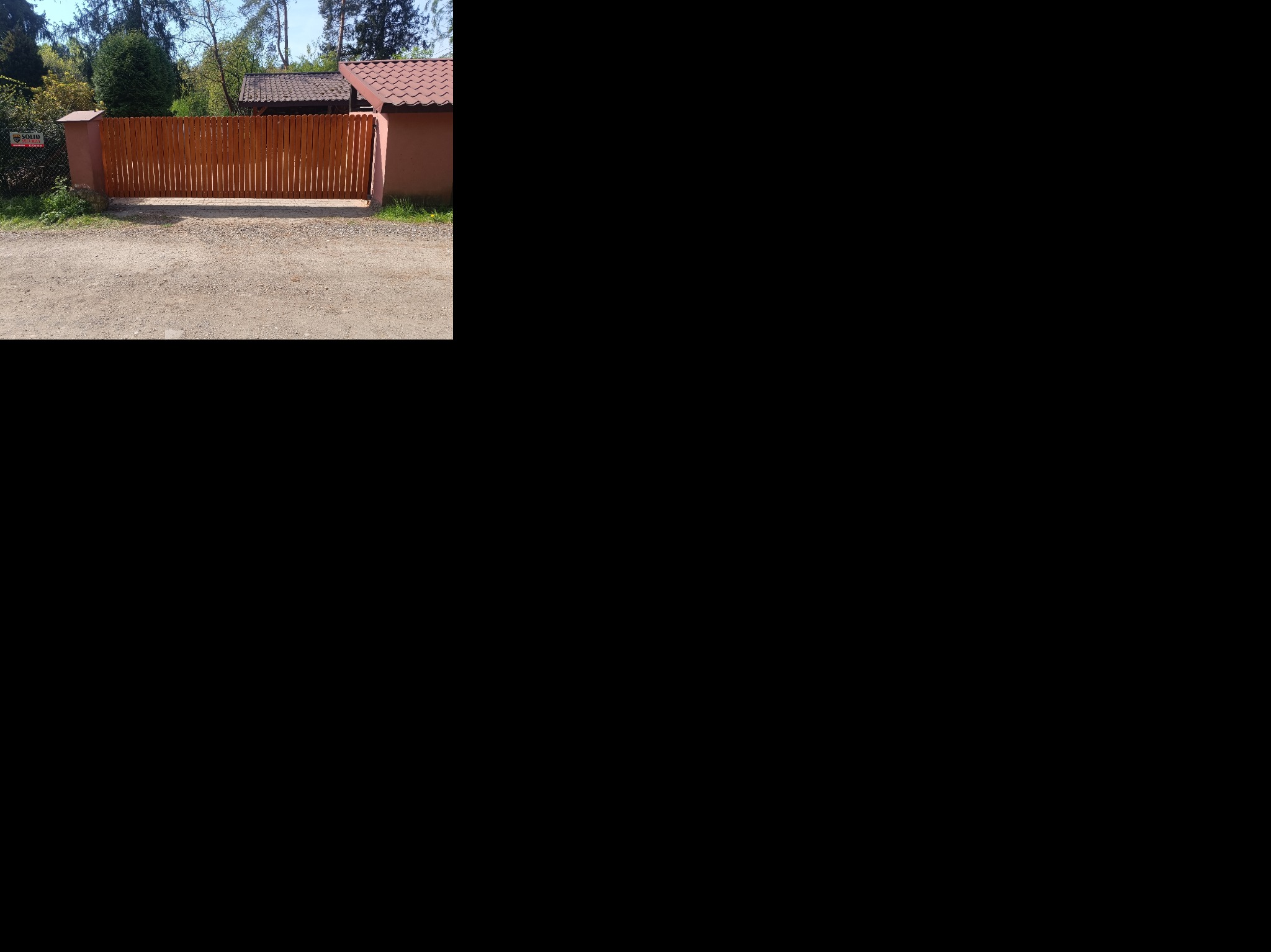 Drewniana brama wjazdowa z pionowych desek, osadzona w murze z cegły, na żwirowym podjeździe. Widoczny fragment dachu budynku w tle.