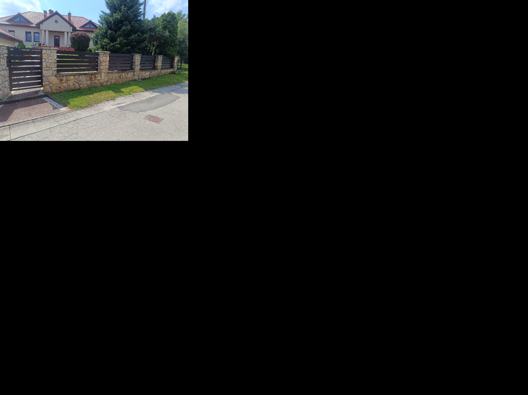 Nowoczesne ogrodzenie z kamienną podmurówką i poziomymi, ciemnymi deskami, brama wjazdowa, dom w tle, trawnik i chodnik. Ujęcie z poziomu ulicy.