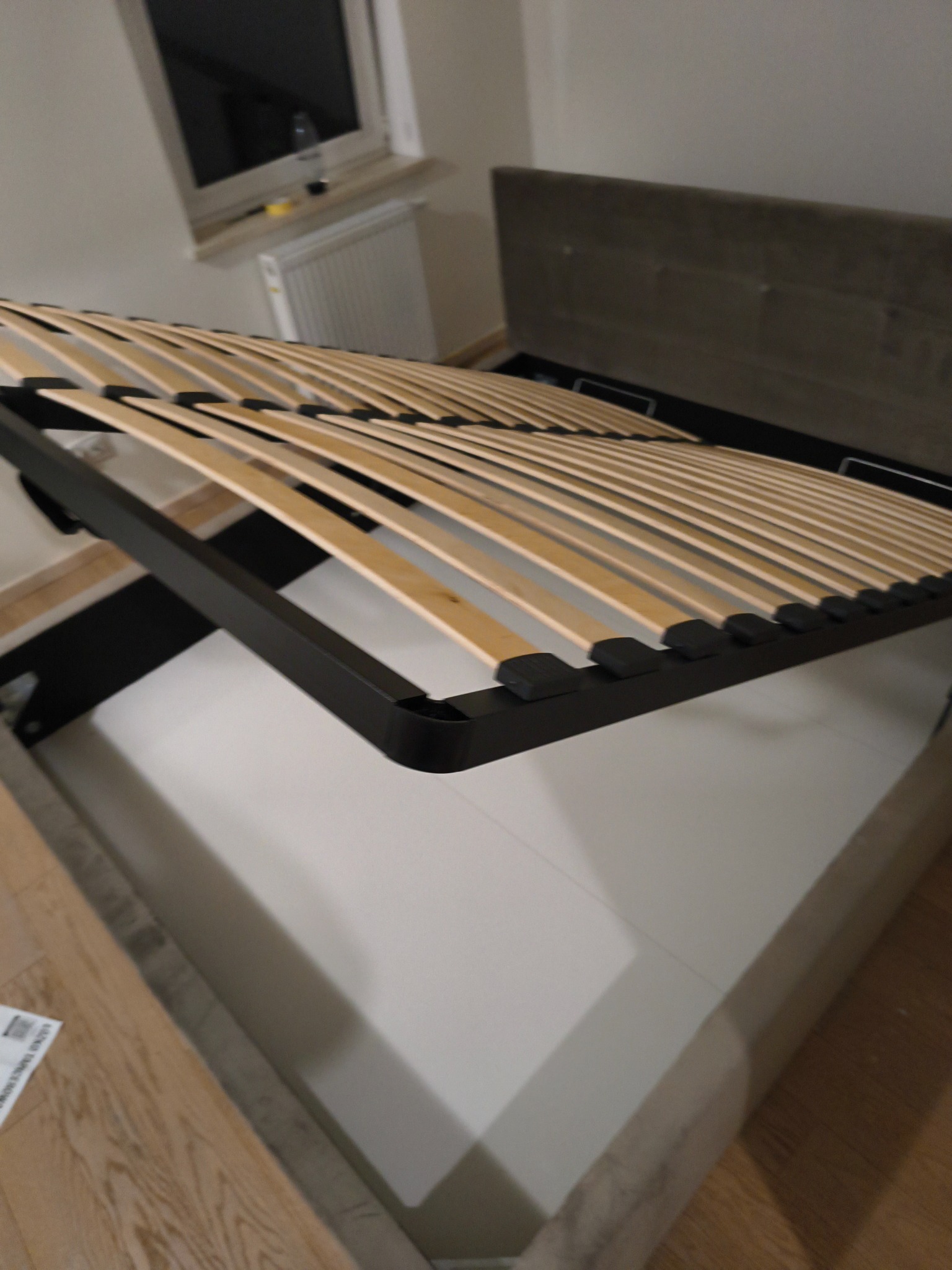 Rozłożony stelaż łóżka z pojemnikiem, widoczne drewniane listwy i mechanizm podnoszenia. Szara tapicerka ramy łóżka, jasne tło ściany i podłogi.