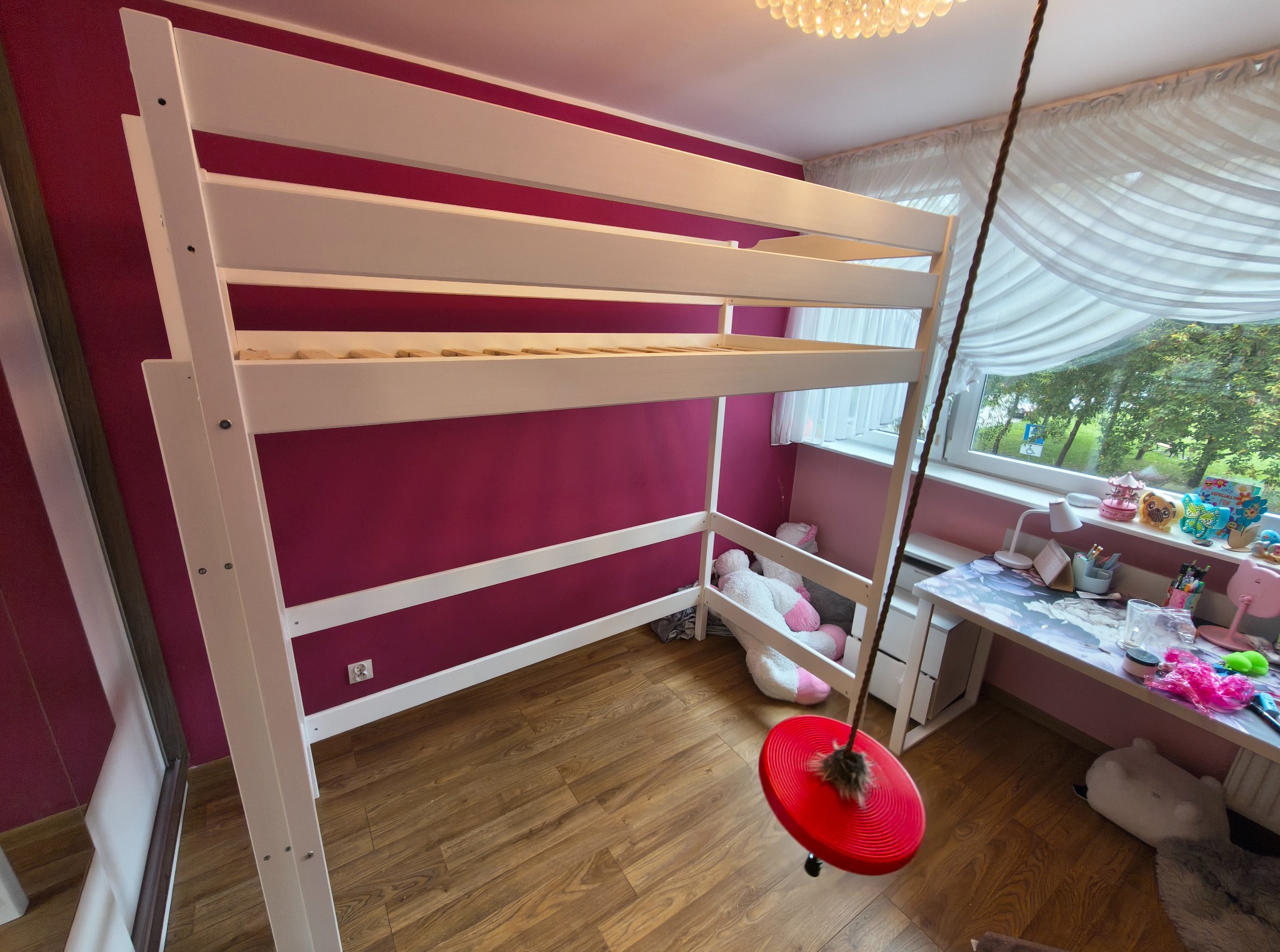 Pokój dziecięcy z białym łóżkiem piętrowym, biurkiem pod oknem i huśtawką na linie. Ściany w kolorze fuksji, drewniana podłoga. Widok na zewnątrz przez okno.