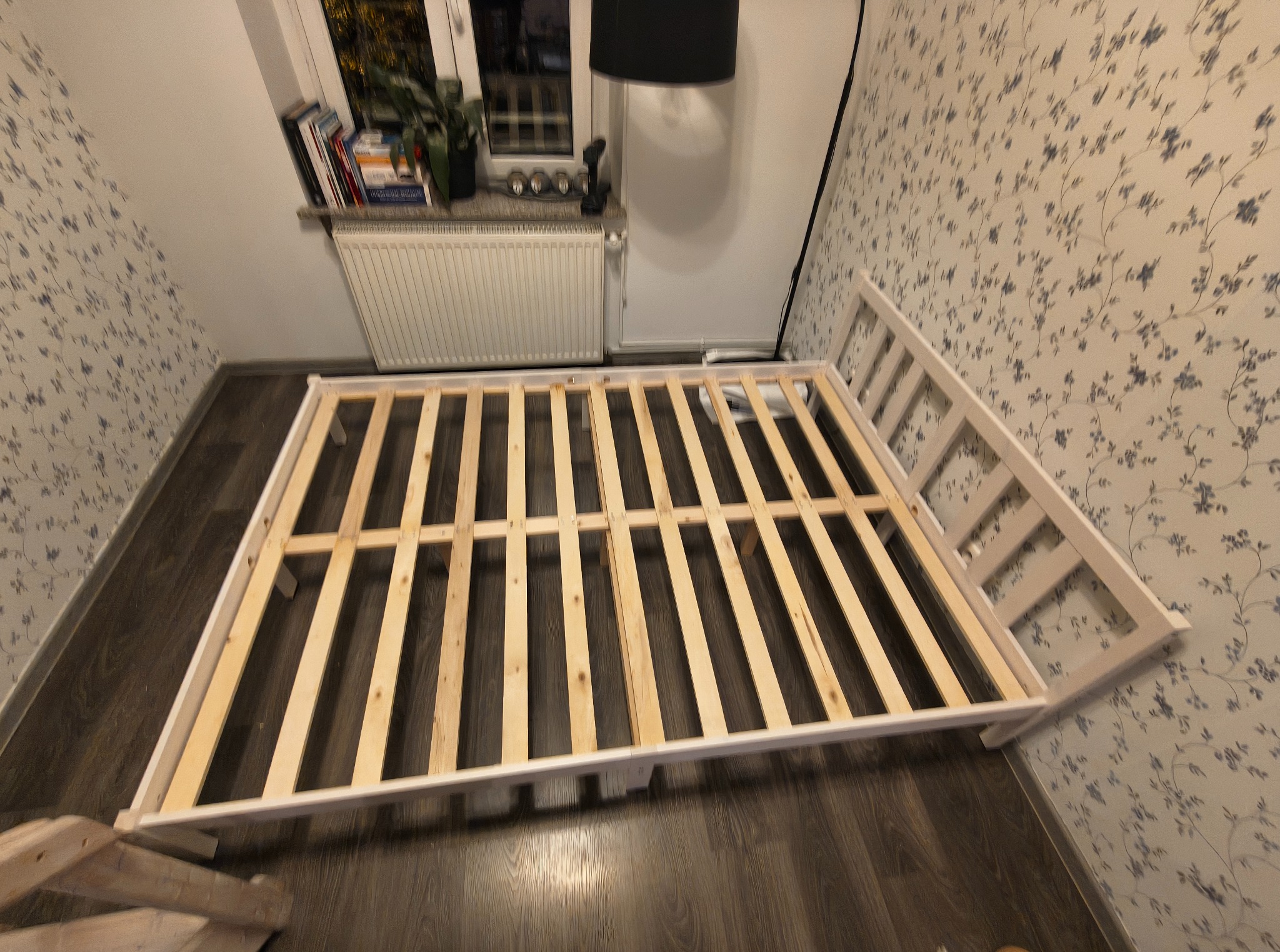Biała rama łóżka z drewnianymi listwami na podłodze, częściowo zmontowana, na tle ściany w kwiaty i okna z roślinami. Widok z góry, ukośny kadr.