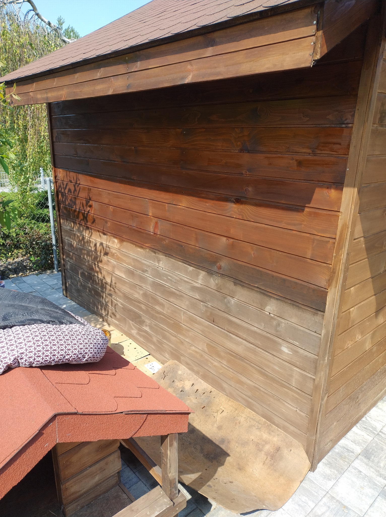 Impregnacja drewnianej altany ogrodowej w trakcie realizacji. Widoczna różnica w kolorze drewna przed i po nałożeniu impregnatu. Częściowo zakryta buda dla psa.