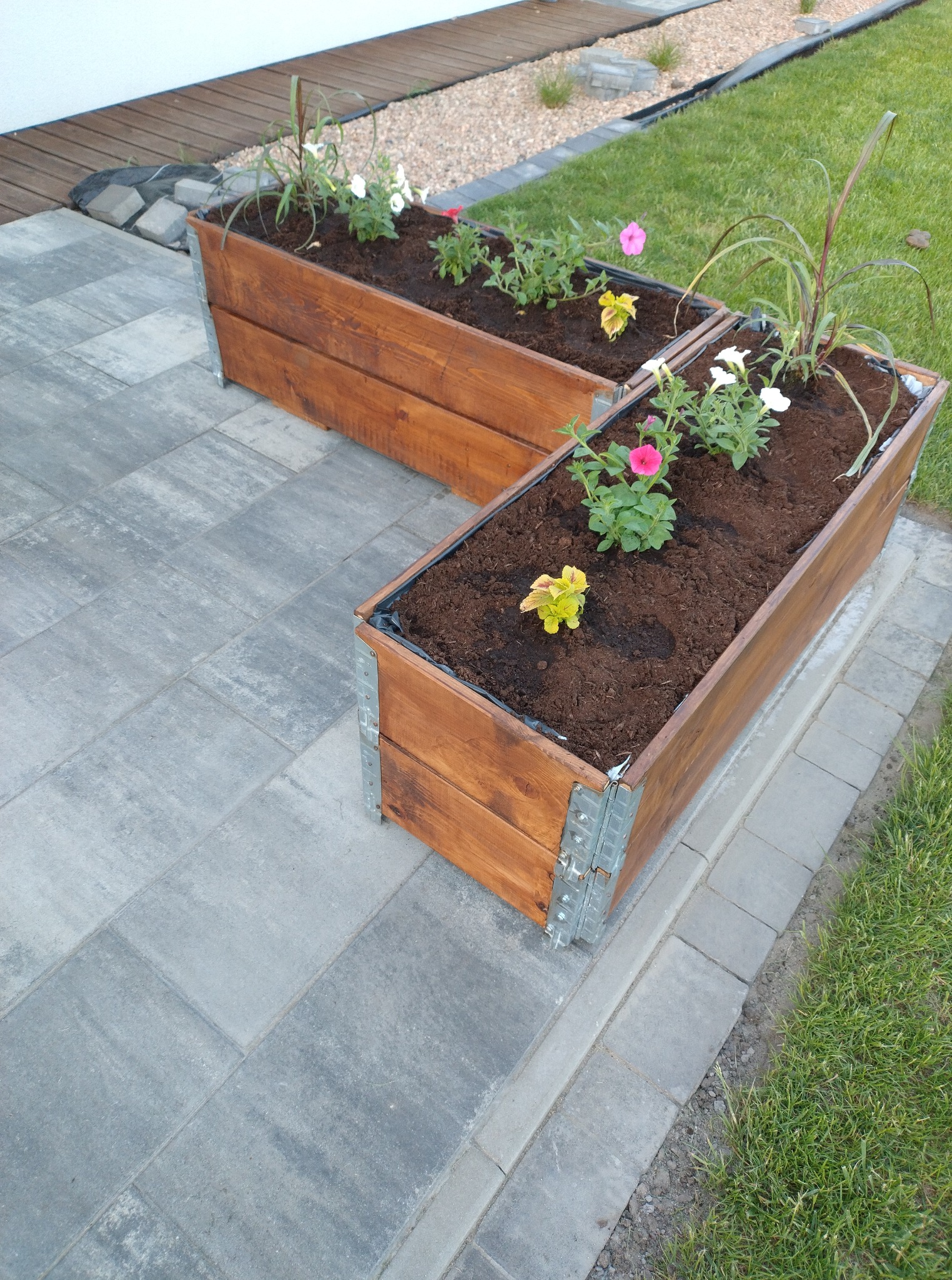 Dwie drewniane skrzynie na kwiaty z metalowymi wzmocnieniami, ustawione na szarym bruku, z widoczną trawą i elementami drewnianej podłogi w tle.