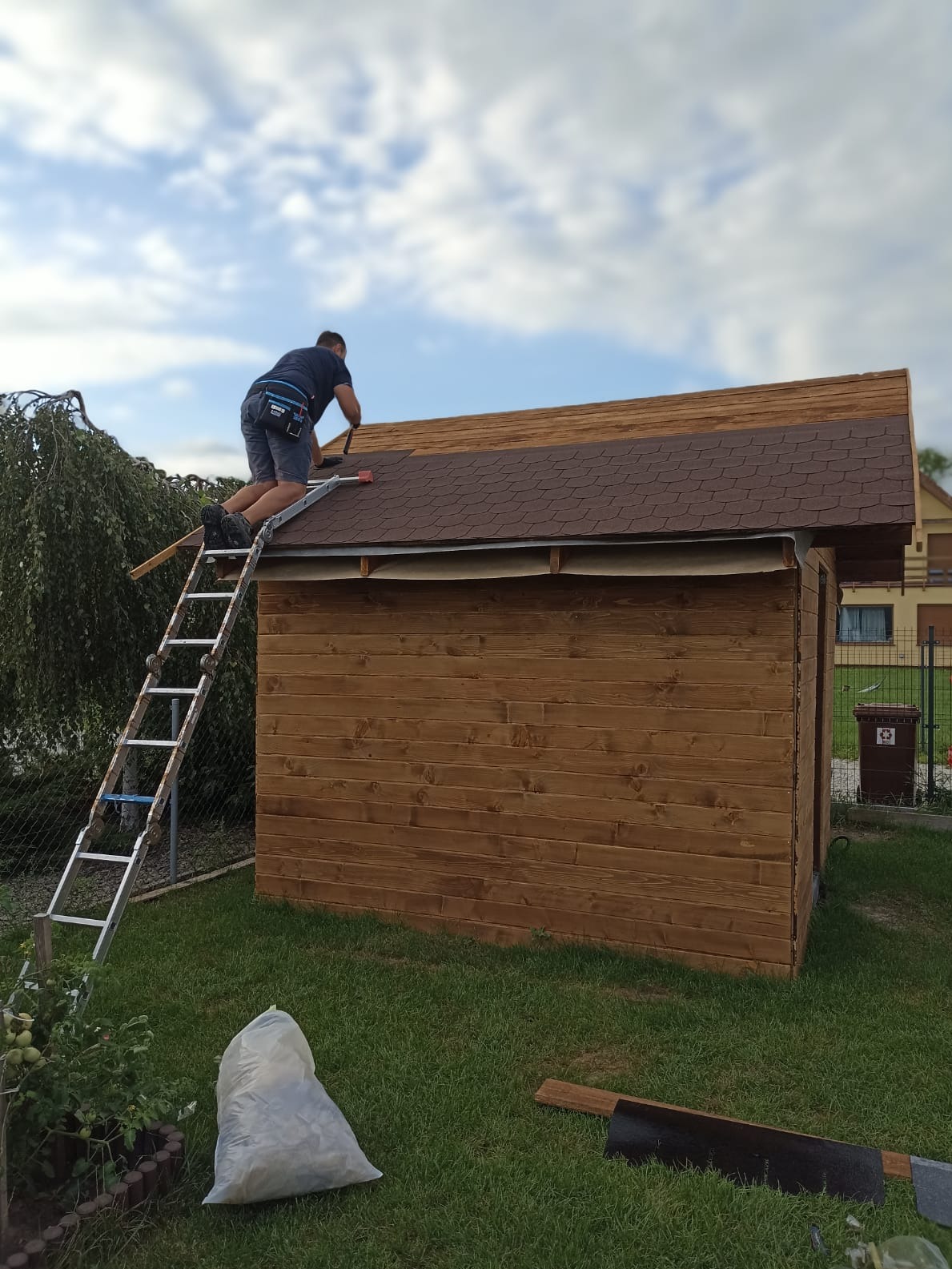 Pracownik na drabinie montuje brązowy gont bitumiczny na dachu drewnianej altany w ogrodzie. Widoczny worek z odpadami i narzędzia.