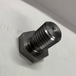 MAGMA Tech - Metalowy, gwintowany element z sześciokątną podstawą, leżący na białym tle. Widoczne ślady obróbki i połysk metalu.