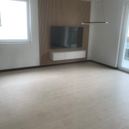 GL wykończenia - Jasne panele podłogowe w nowoczesnym salonie z drewnianą ścianą TV i minimalistyczną lampą wiszącą. Widok na okno i drzwi balkonowe.