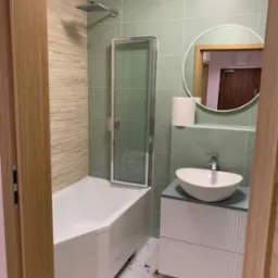 Nowoczesna łazienka z zielonymi płytkami, białą wanną z parawanem, okrągłym lustrem i umywalką nablatową na szafce z ryflowanym frontem. Podłoga w stylu lastryko.