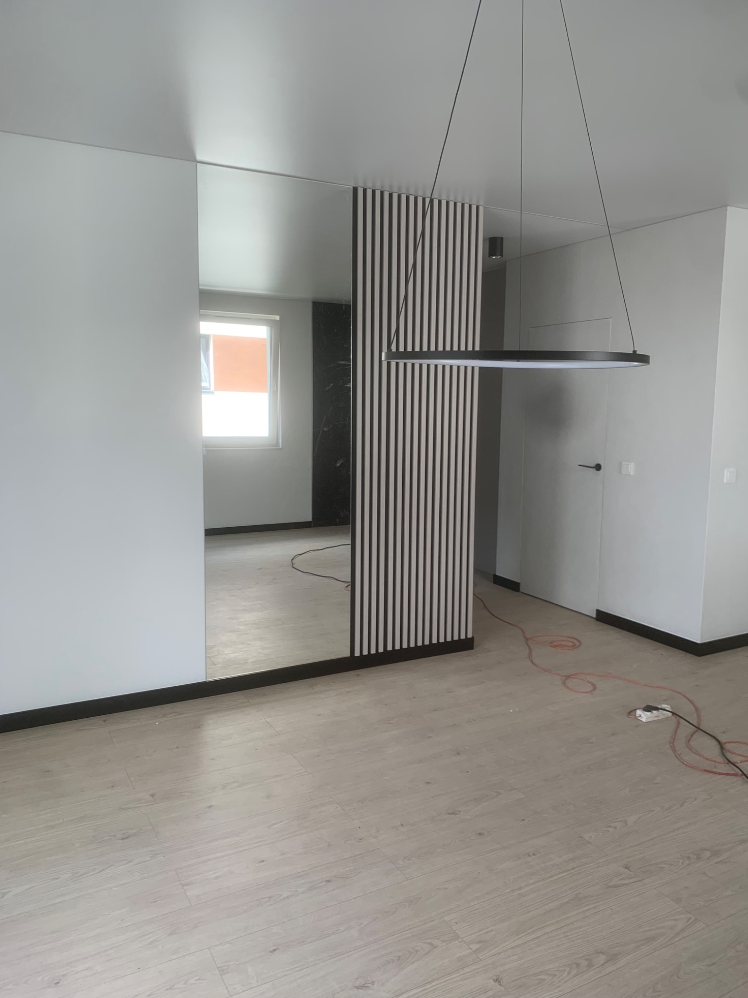 Nowoczesne wnętrze z drewnianą ścianką działową, lustrem i okrągłą lampą wiszącą. Jasne ściany i podłoga, minimalistyczny design. Widoczne gniazdko elektryczne na podłodze.