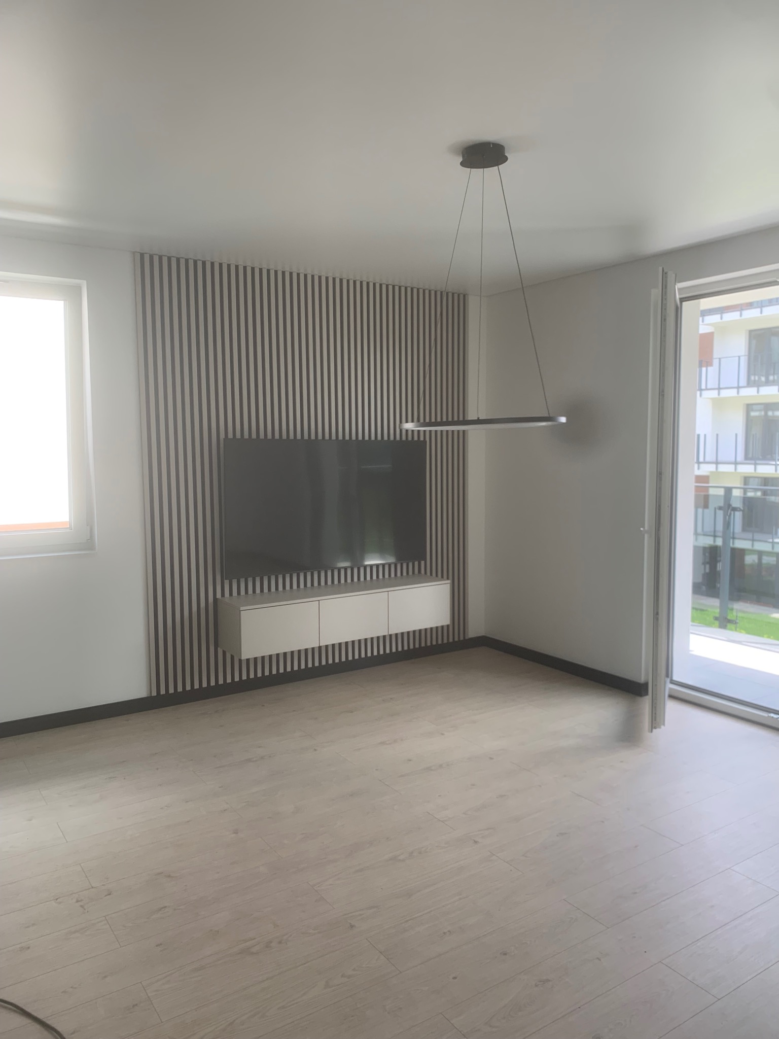 Nowoczesny salon z drewnianą ścianą z lameli, podwieszaną szafką RTV i minimalistyczną lampą. Jasna podłoga i duże okno doświetlają wnętrze.