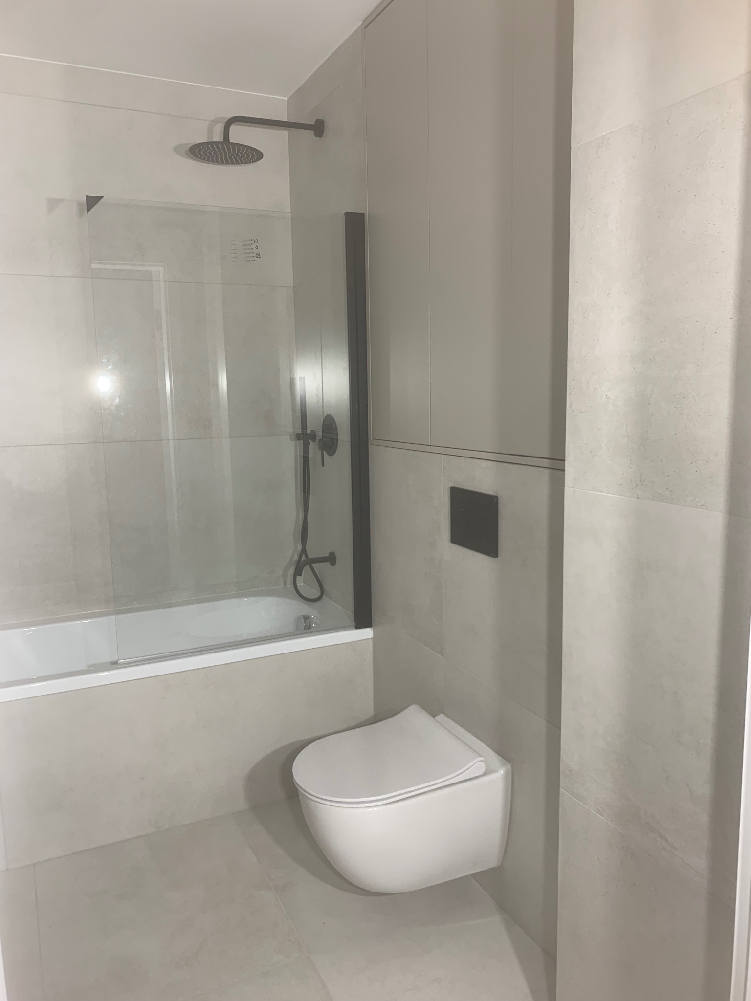 Nowoczesna łazienka w jasnych odcieniach szarości z kabiną prysznicową typu walk-in, podwieszaną toaletą i deszczownicą. Minimalistyczny design, płytki imitujące kamień.