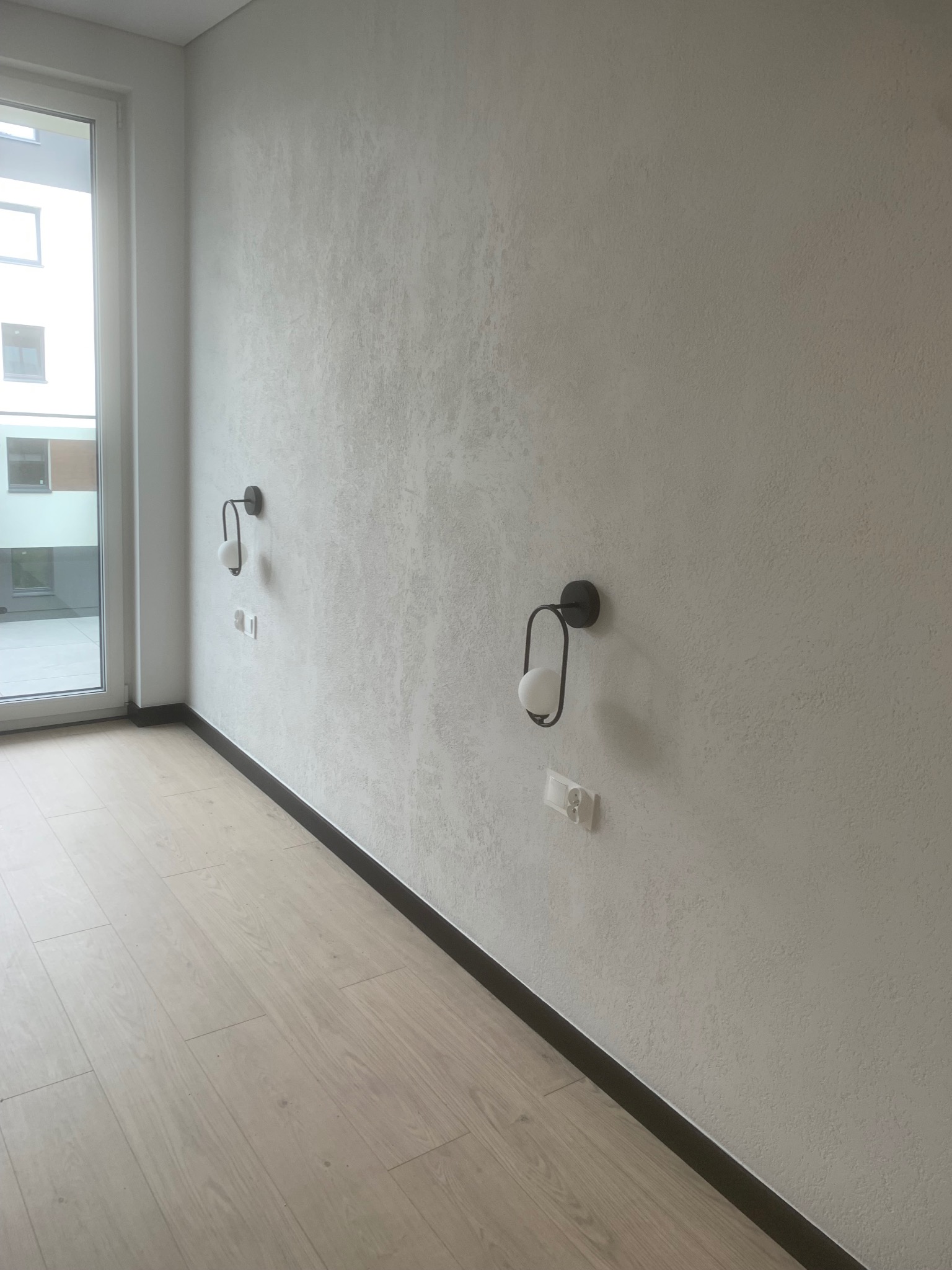 Jasne wnętrze po wykończeniu: drewniana podłoga, ściana z fakturą, nowoczesne lampy ścienne i listwy przypodłogowe. Widok na okno balkonowe.