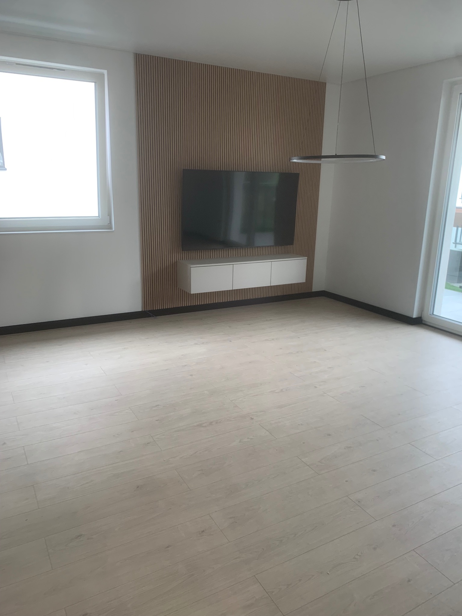 Jasne panele podłogowe w nowoczesnym salonie z drewnianą ścianą TV i minimalistyczną lampą wiszącą. Widok na okno i drzwi balkonowe.