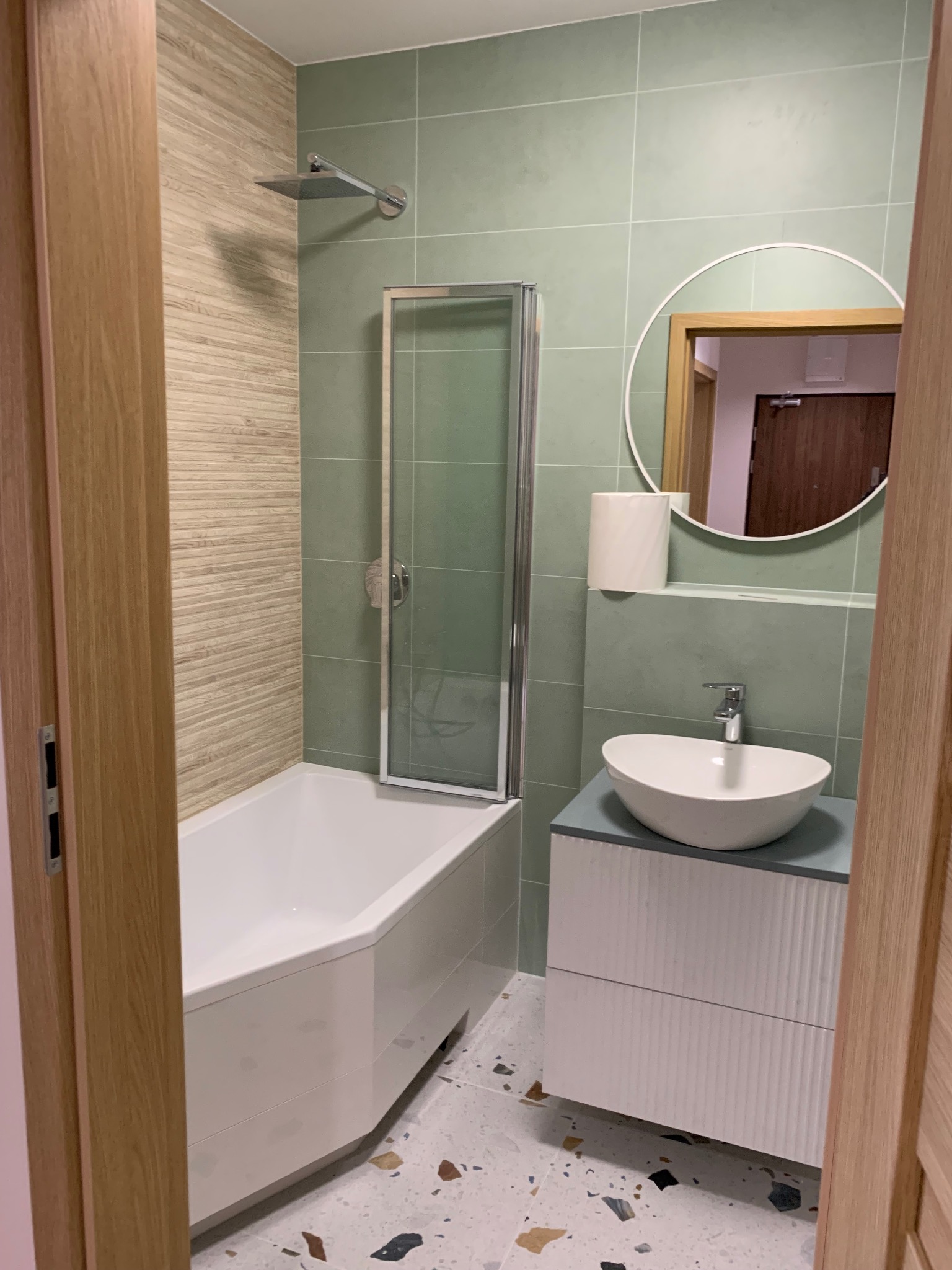 Nowoczesna łazienka z zielonymi płytkami, białą wanną z parawanem, okrągłym lustrem i umywalką nablatową na szafce z ryflowanym frontem. Podłoga w stylu lastryko.