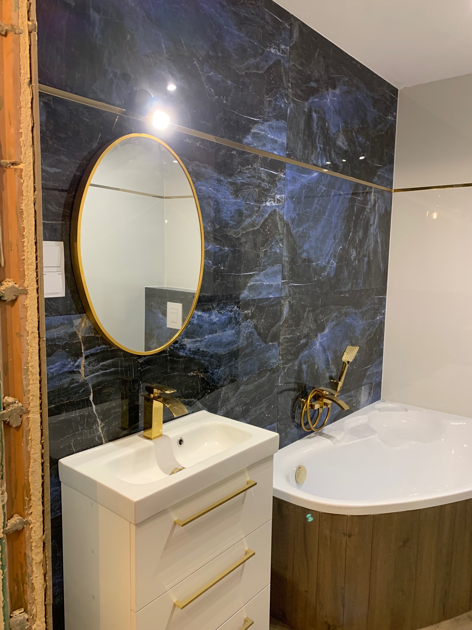 Wykończona łazienka z umywalką na szafce, owalnym lustrem w złotej ramie i wanną narożną. Na ścianach płytki imitujące marmur z niebieskimi żyłami i złote akcenty.