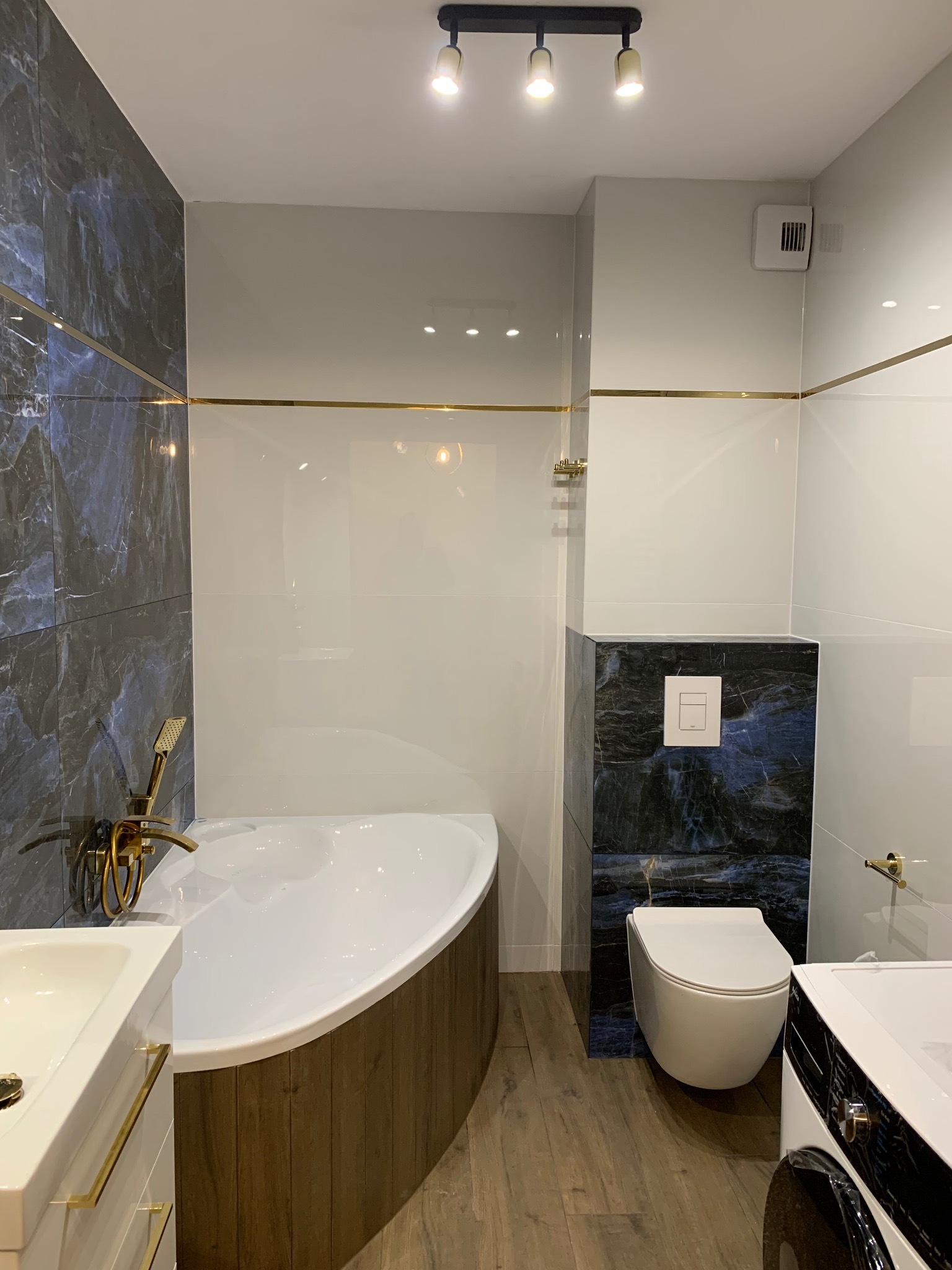 Nowoczesna łazienka z wanną obudowaną drewnem, marmurowymi akcentami i złotymi detalami armatury, oświetlona punktowo.