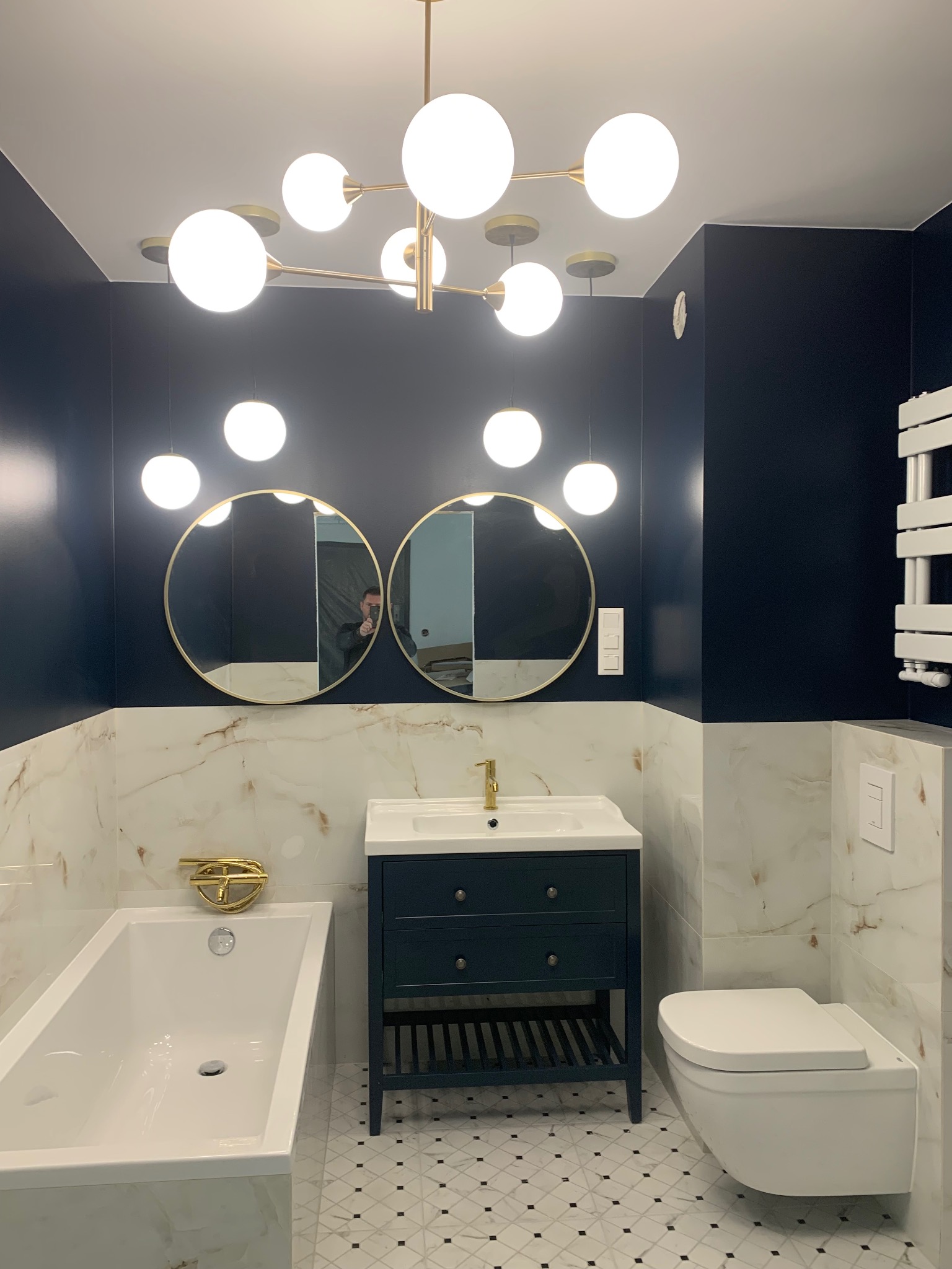 Elegancka łazienka w odcieniach granatu i bieli z marmurowymi płytkami, okrągłymi lustrami w złotych ramach i nowoczesnym oświetleniem.