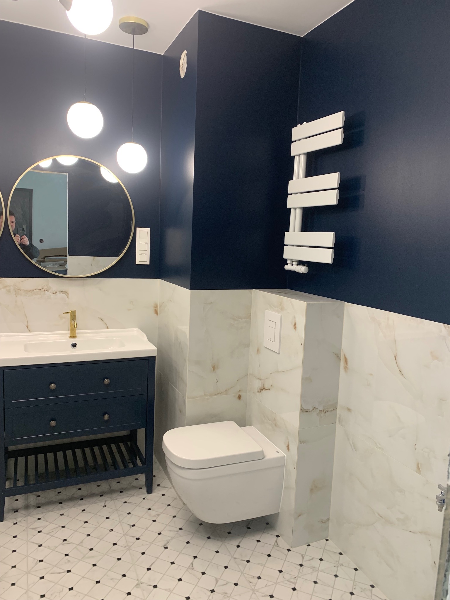 Elegancka łazienka w odcieniach granatu i bieli. Umywalka z szafką, okrągłe lustro, podwieszana toaleta i grzejnik drabinkowy. Marmurowe płytki i mozaika na podłodze.