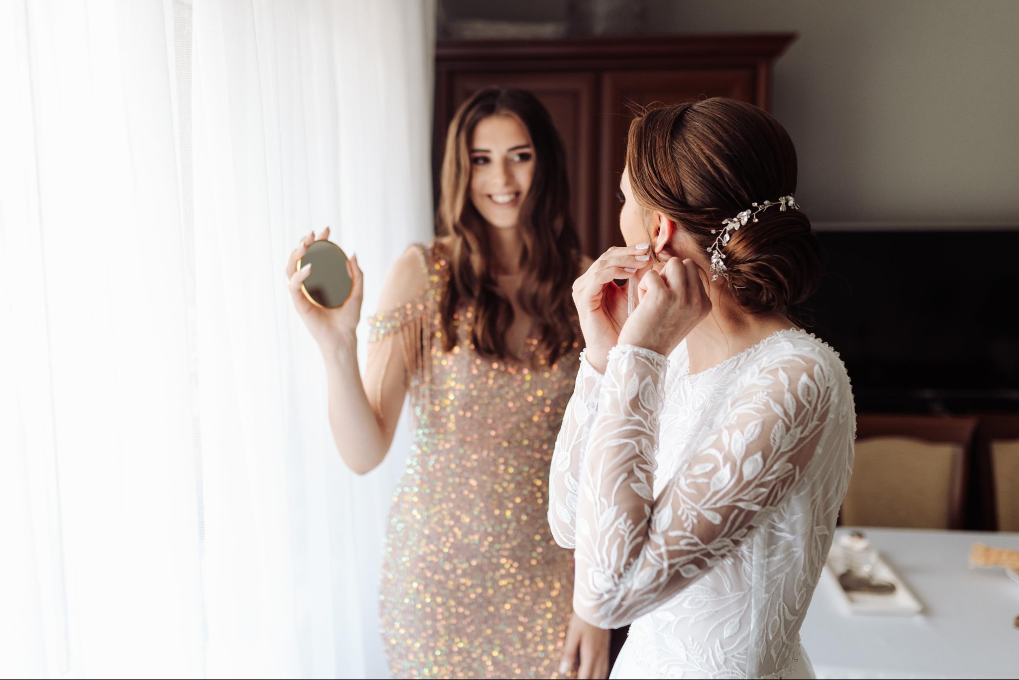 Panna młoda w białej sukni z koronkowymi rękawami zakłada kolczyk, obok uśmiechnięta druhna trzyma lusterko na tle okna.