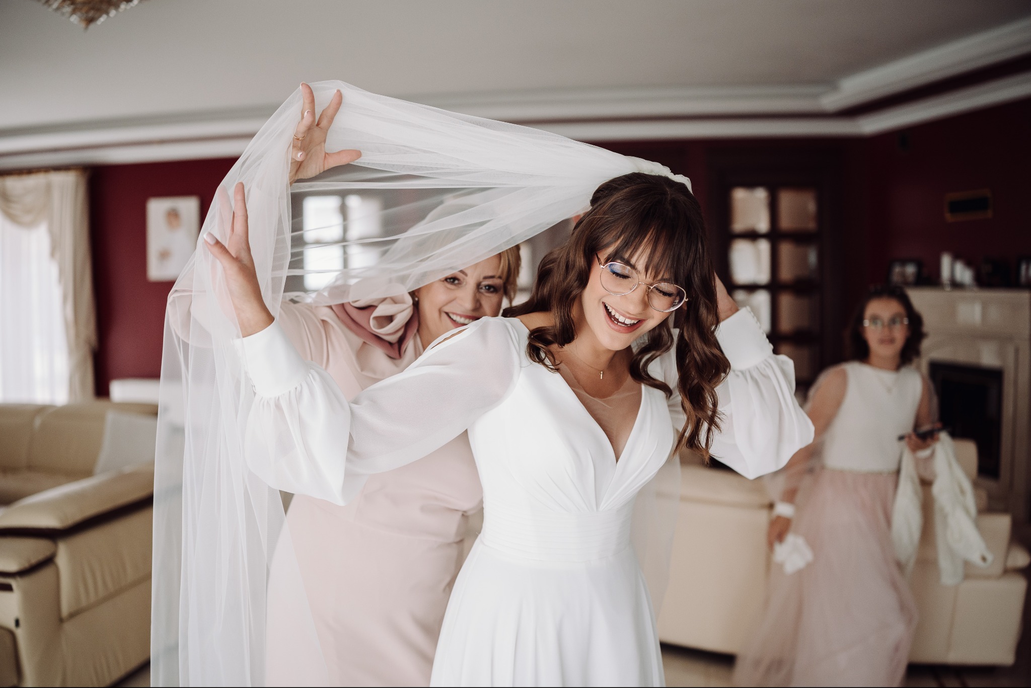 Uśmiechnięta panna młoda w okularach, w białej sukni, podczas zakładania welonu przez kobietę w różowej sukience. W tle druhny i wnętrze domu.