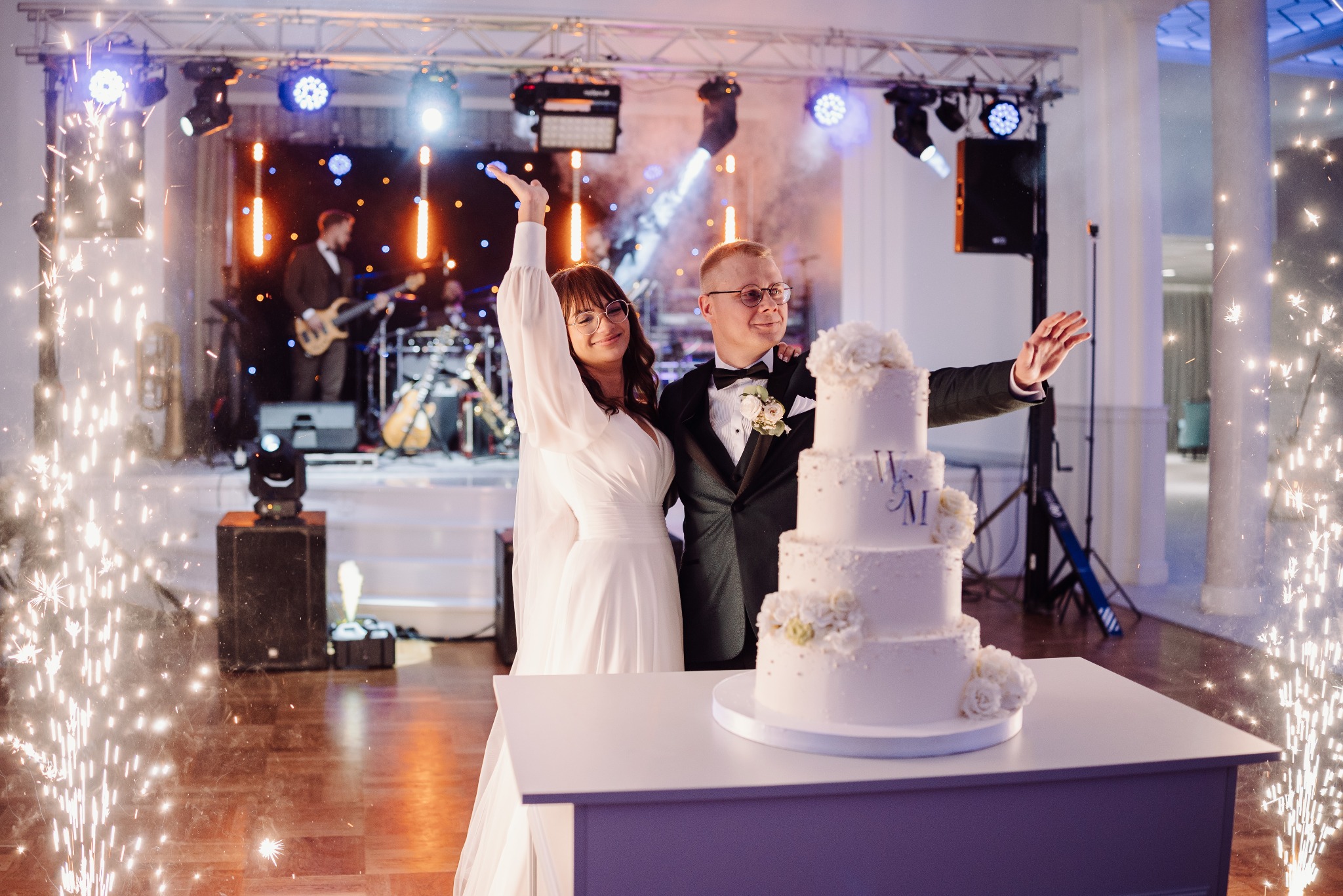 Uśmiechnięta para młoda w eleganckich strojach pozuje przy wielopiętrowym torcie weselnym, otoczona fontannami iskier na tle sceny z zespołem muzycznym.