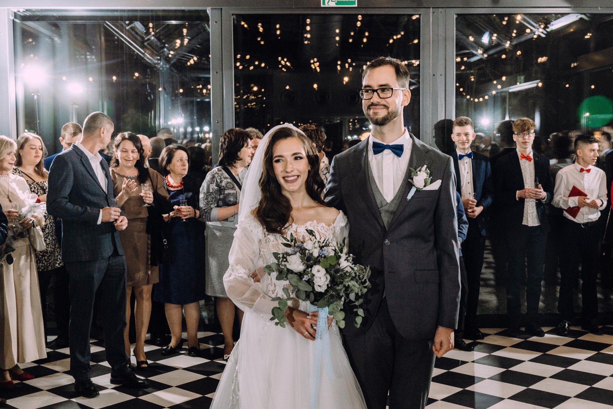 Uśmiechnięta para młoda w otoczeniu gości weselnych na tle czarno-białej podłogi. Panna młoda trzyma bukiet. W tle widoczne dekoracje świetlne.
