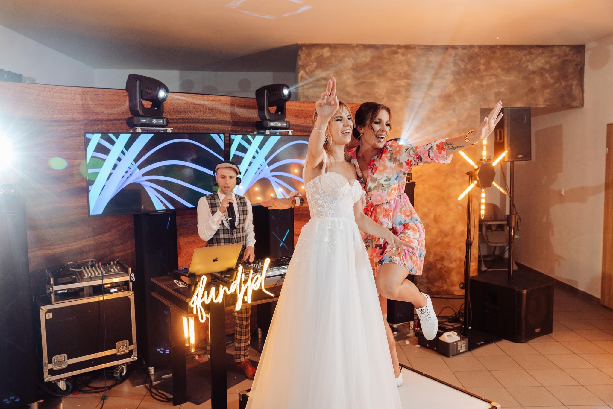 Uśmiechnięta panna młoda w białej sukni i kobieta w kwiecistej sukience tańczą na weselu. W tle DJ z mikrofonem i sprzęt muzyczny.
