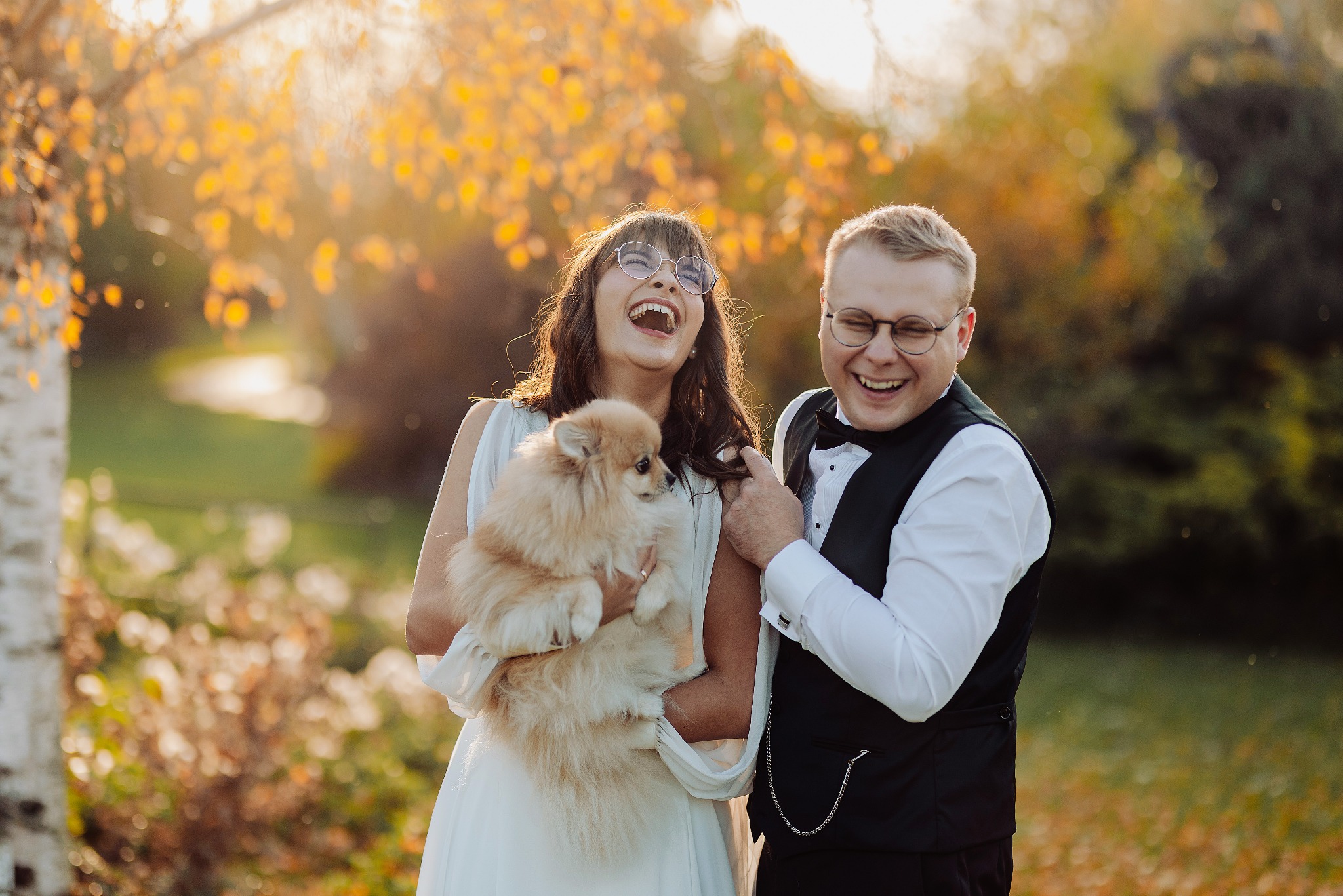 Uśmiechnięta para młoda w okularach, trzymająca szpica miniaturowego na tle jesiennego parku. Pan młody w czarnej kamizelce i muszce, panna młoda w białej sukni.