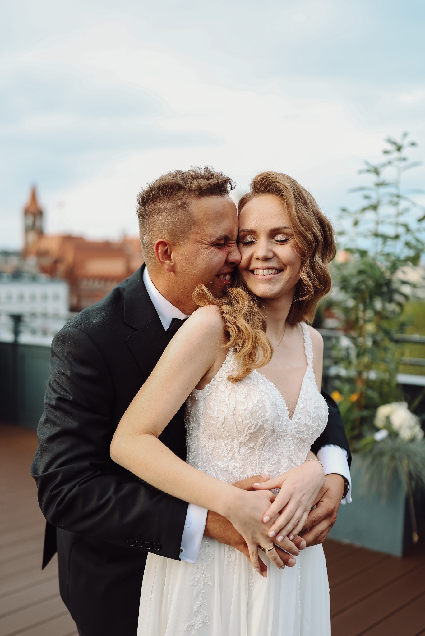 Uśmiechnięta para młoda w Kościanie, w objęciach na tle miejskiej zabudowy. Panna młoda w koronkowej sukni, pan młody w czarnym garniturze. Romantyczna sesja.