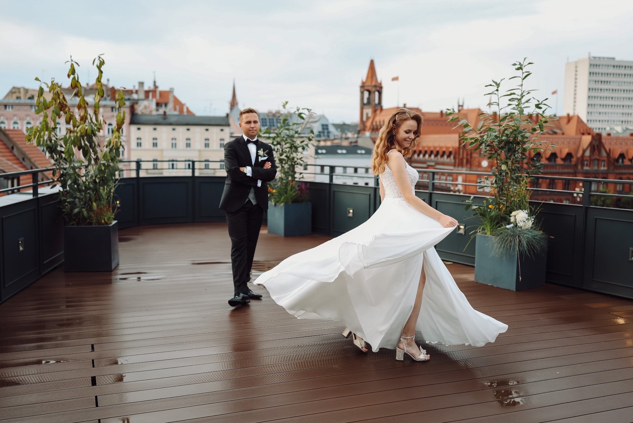 Para młoda na tle miejskiej panoramy: panna młoda w tańcu, pan młody z założonymi rękami. Ujęcie z góry, widoczny mokry drewniany taras.